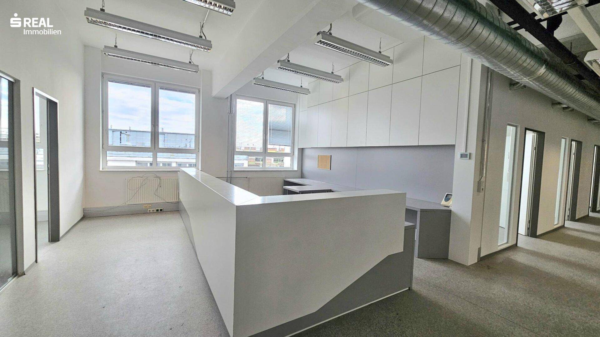 Offener Bürobereich mit Empfangstheke, weißen Schränken und großen Fenstern.