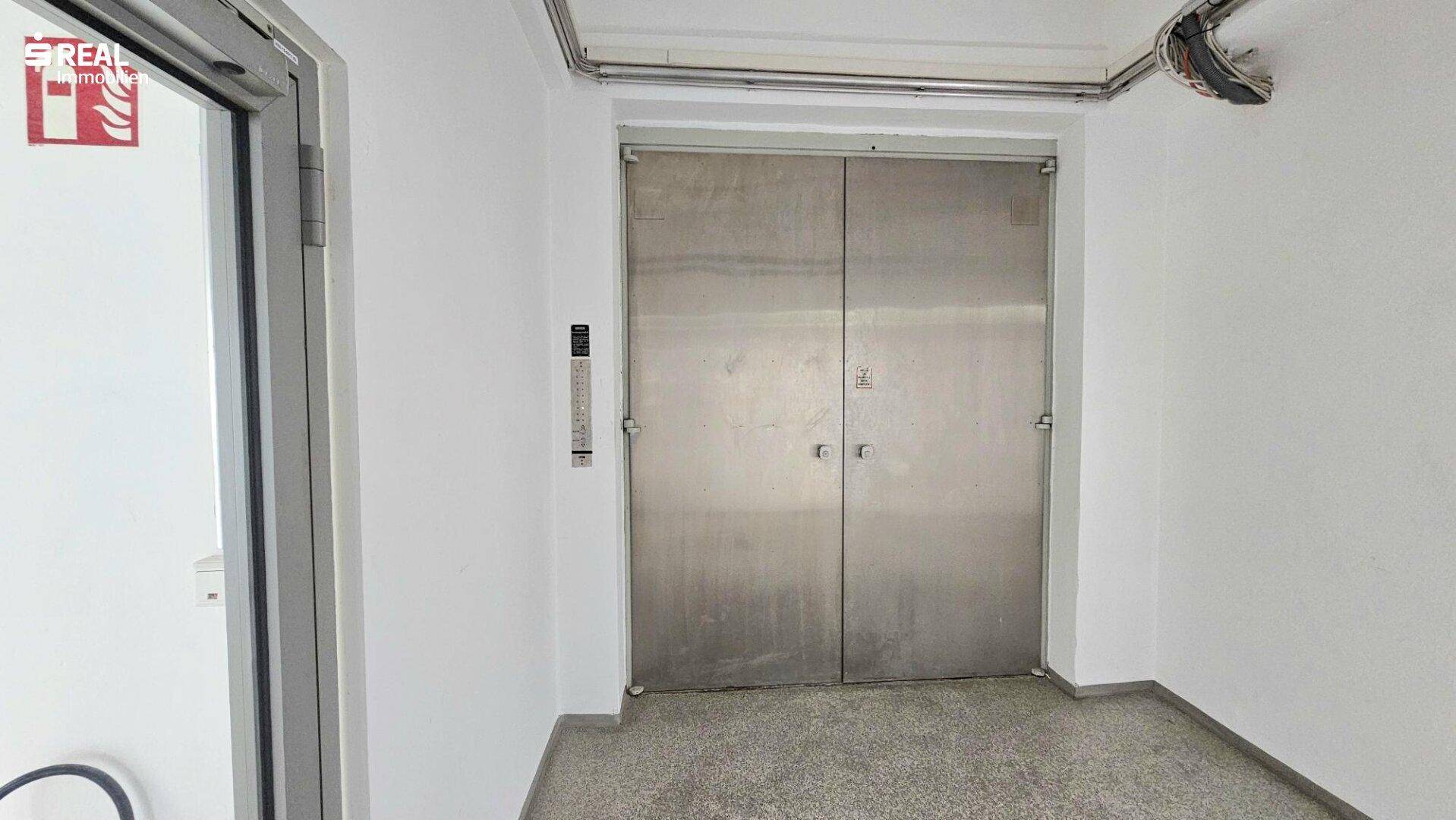 Flur mit großen Metalltüren, die zu einem Lastenaufzug oder Lagerbereich führen.