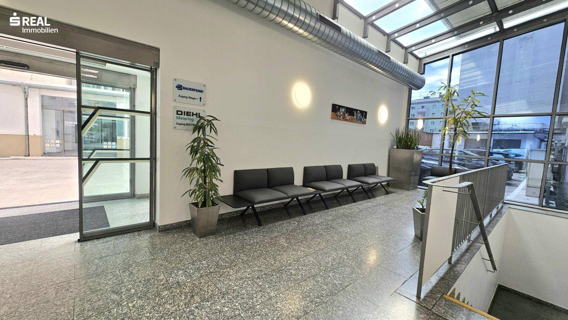 Wartebereich in einer modernen Lobby mit Sitzgelegenheiten, Pflanzen und großen Fenstern.