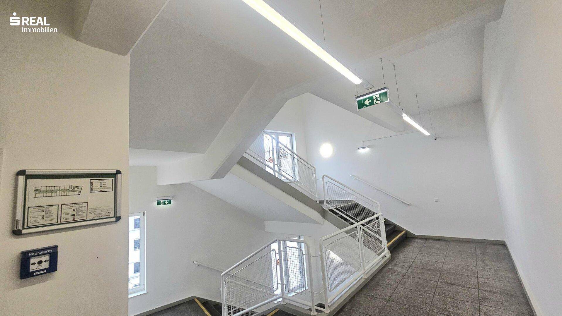 Geräumiges Treppenhaus mit weißen Wänden, Metallgeländer und Notausgangsschildern.