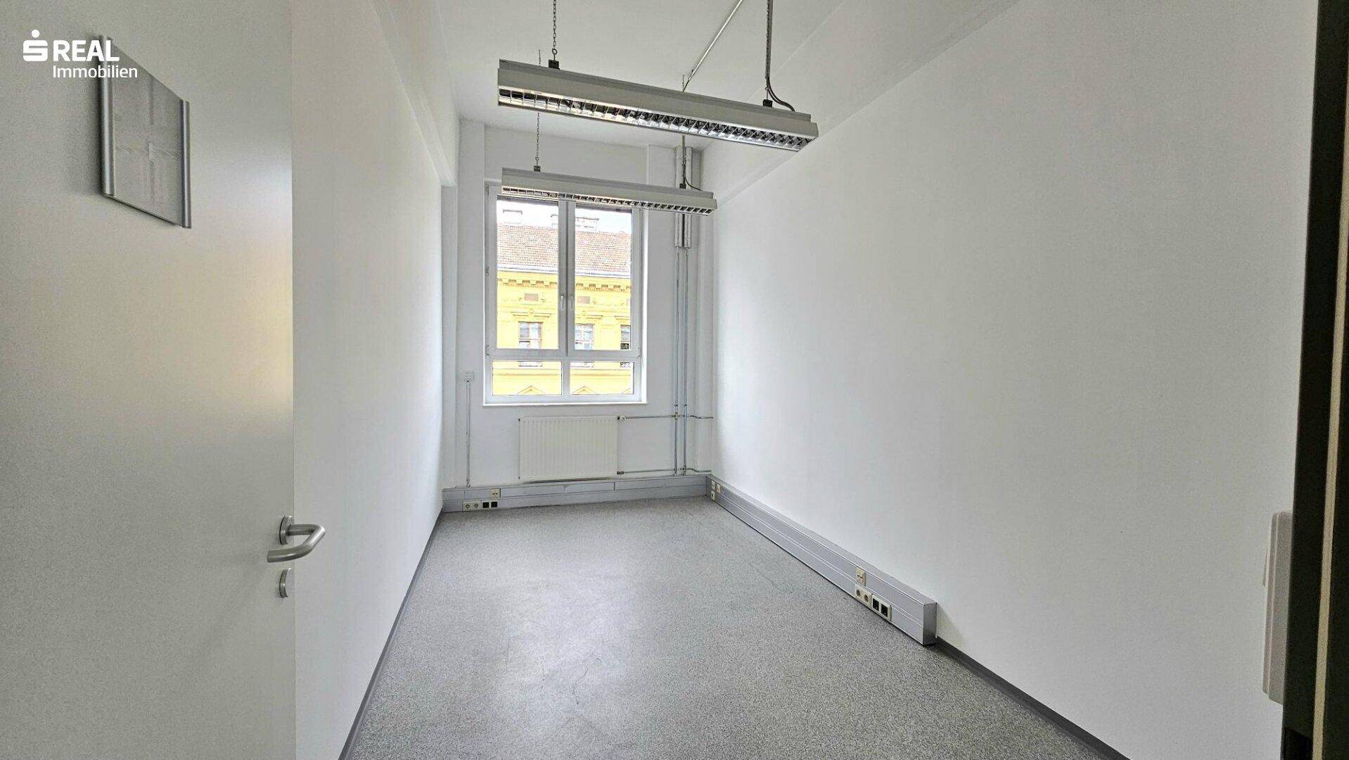 Schmaler Büroraum mit einem Fenster, das Tageslicht hereinlässt, und einem Betonboden.