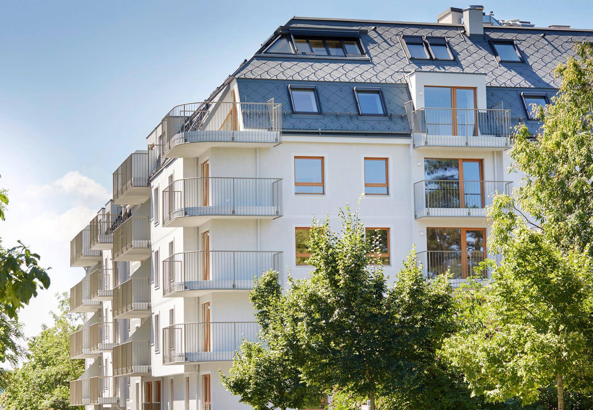 Modernes Mehrfamilienhaus mit Balkonen, Mansarddach und umgebenden Bäumen.