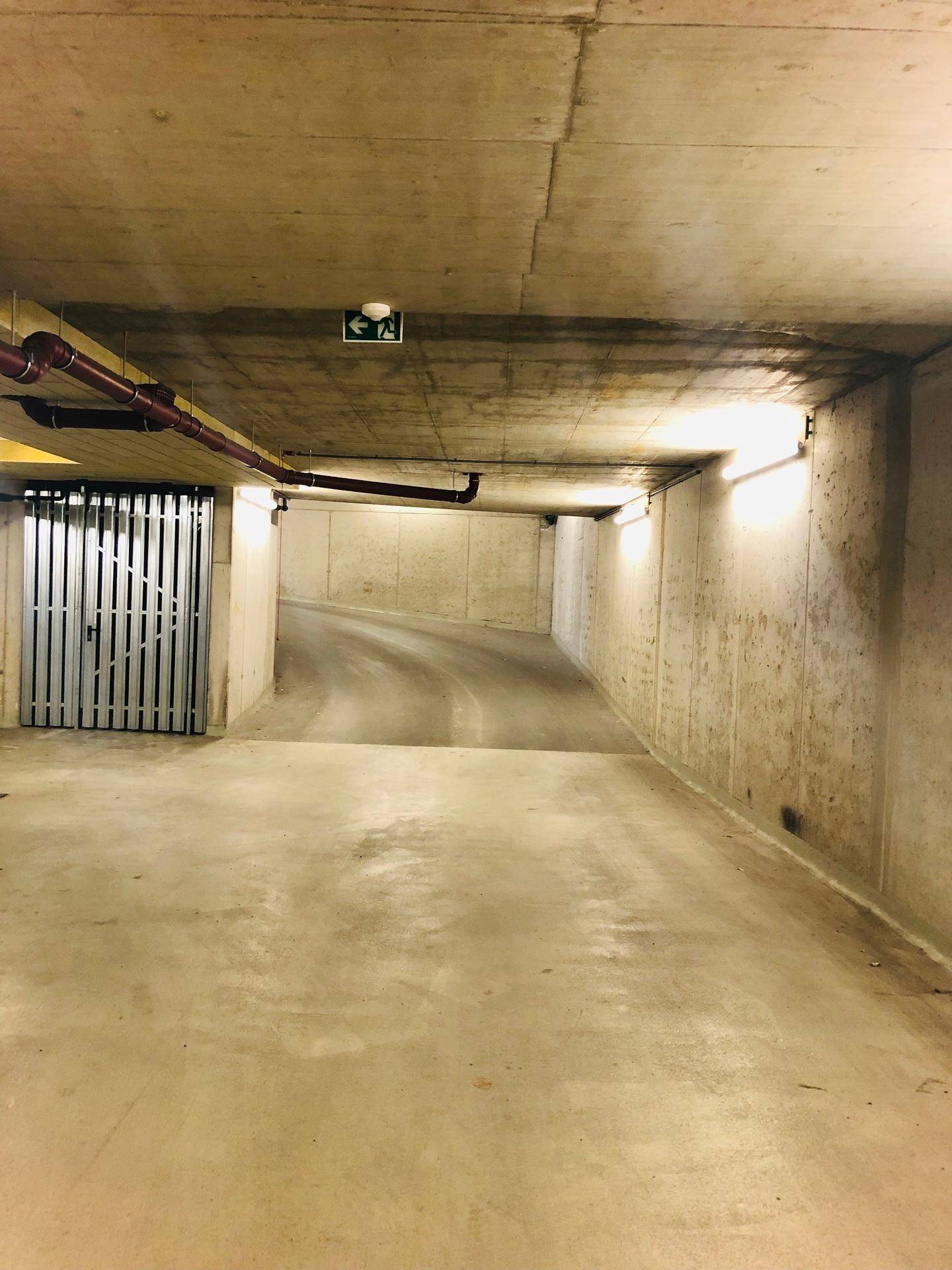 Zufahrtsrampe zur Tiefgarage mit Betonwänden und einer Metalltür im Hintergrund, gut beleuchtet.