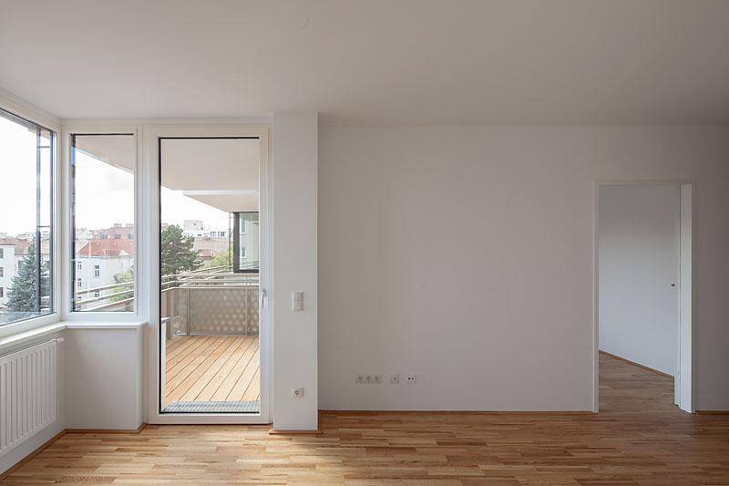 Heller, leerer Raum mit Holzboden und großem Fenster, das Zugang zu einem Balkon bietet.