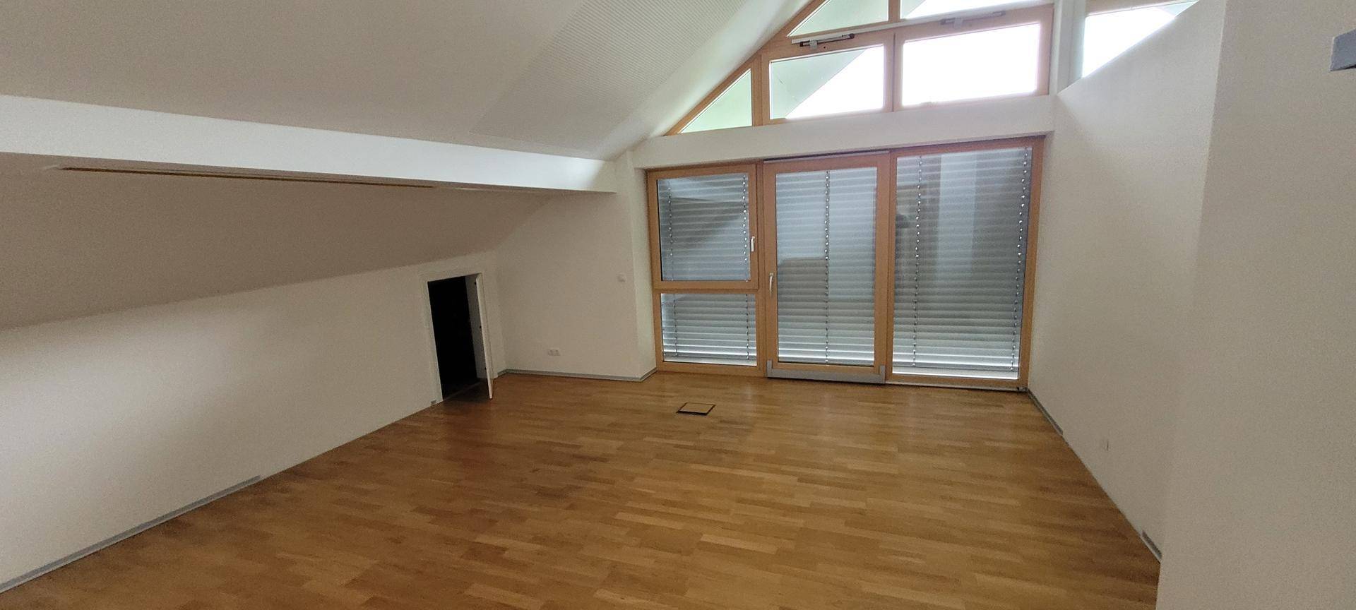 Geräumiges Dachgeschossbüro mit Holzboden, hohen Decken und großen Fenstern mit Jalousien.