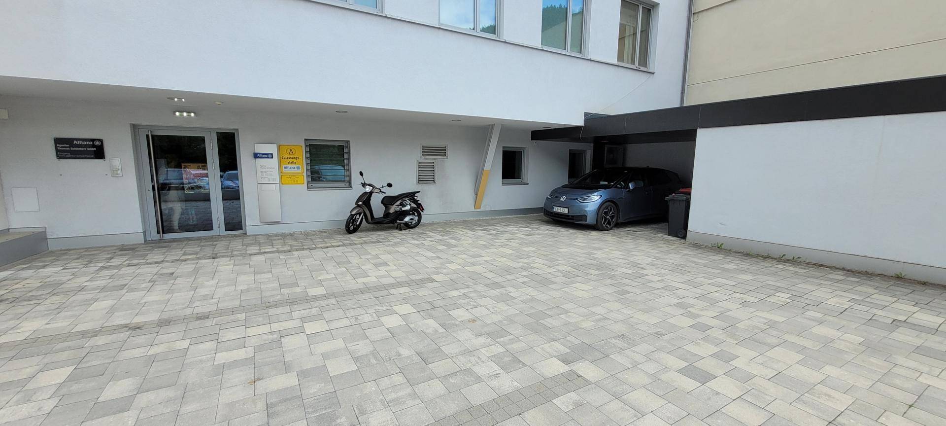 Gepflasterter Vorplatz mit Parkplätzen und einem überdachten Bereich für ein Auto.
