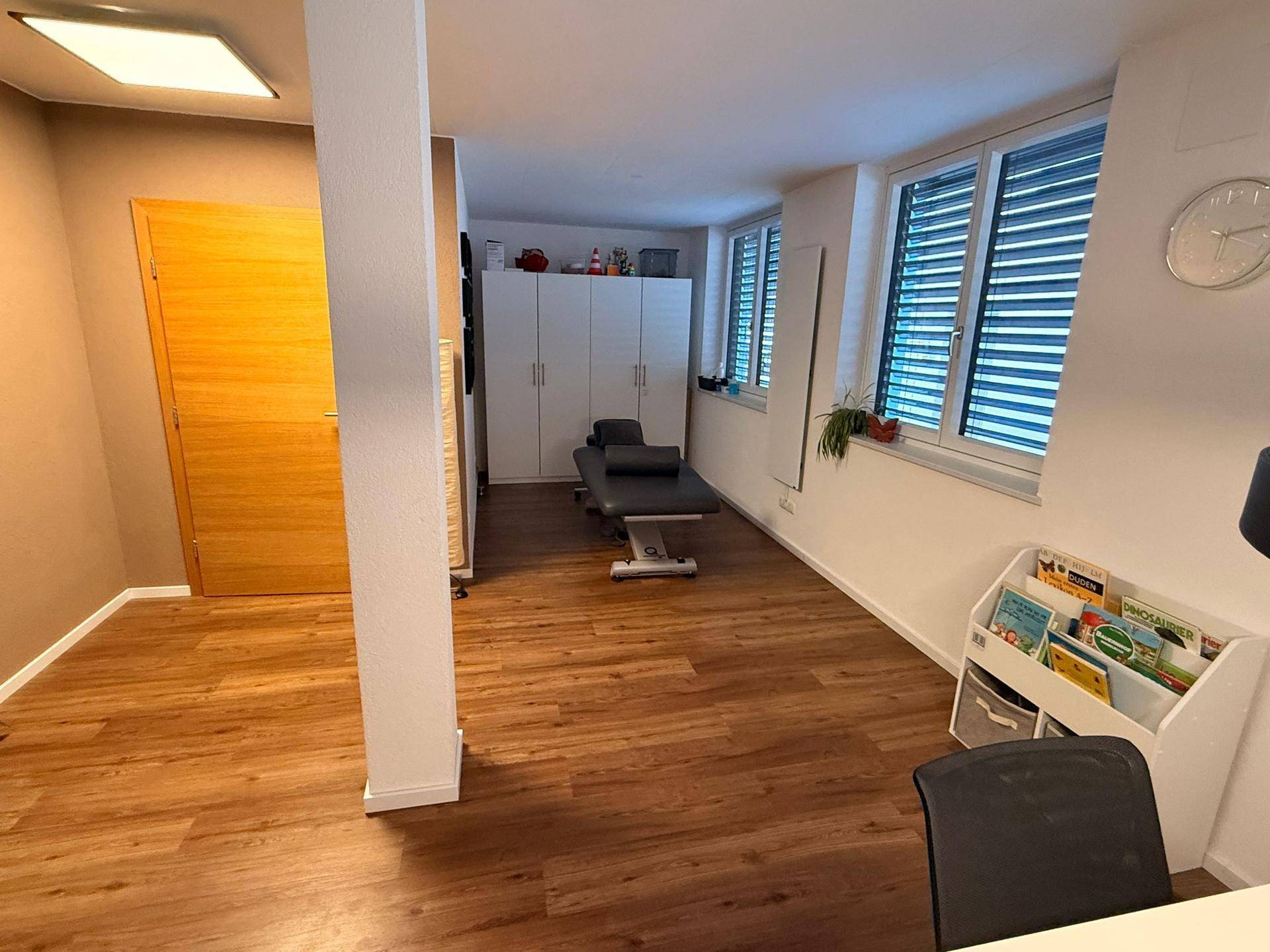 Heller Mehrzweckraum mit Holzboden, Behandlungsliege und weißen Schränken, ideal für therapeutische Zwecke.