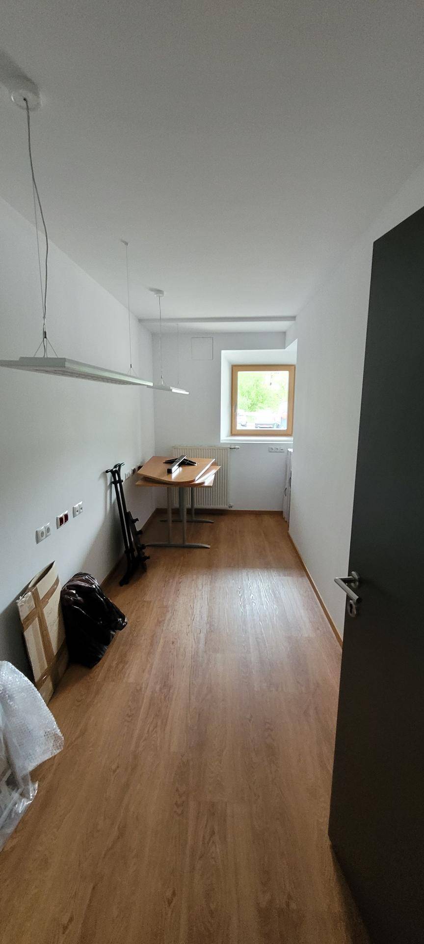 Schmaler Büroraum mit Holzboden, einem kleinen Tisch und einem Fenster.