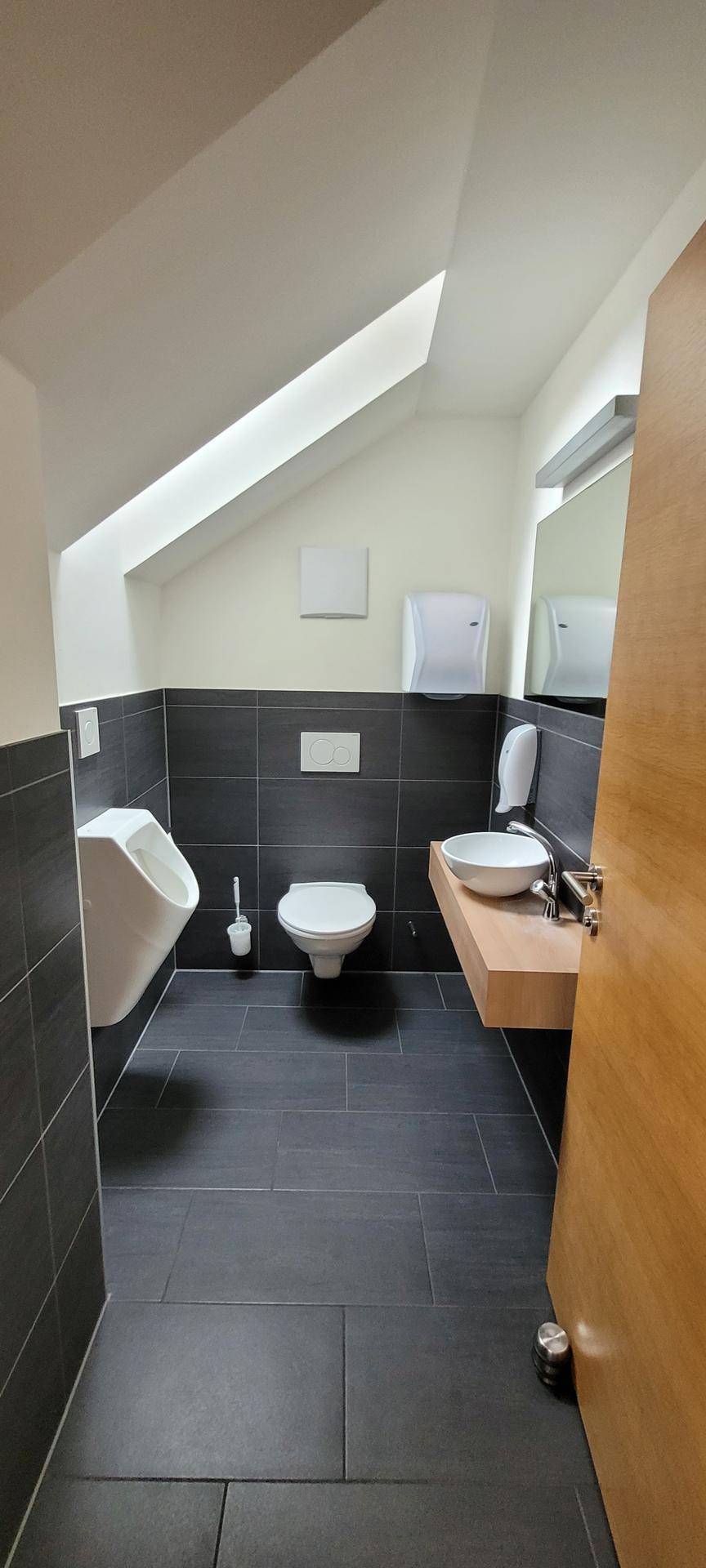 Modernes Badezimmer mit dunklen Fliesen, Urinal, Toilette und Waschbecken unter Dachschräge.