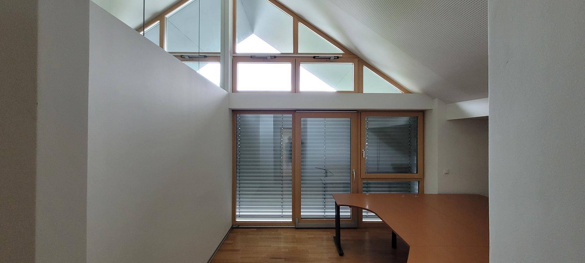Helles Dachgeschossbüro mit Holzboden, hohen Decken und großen Fenstern mit Jalousien.