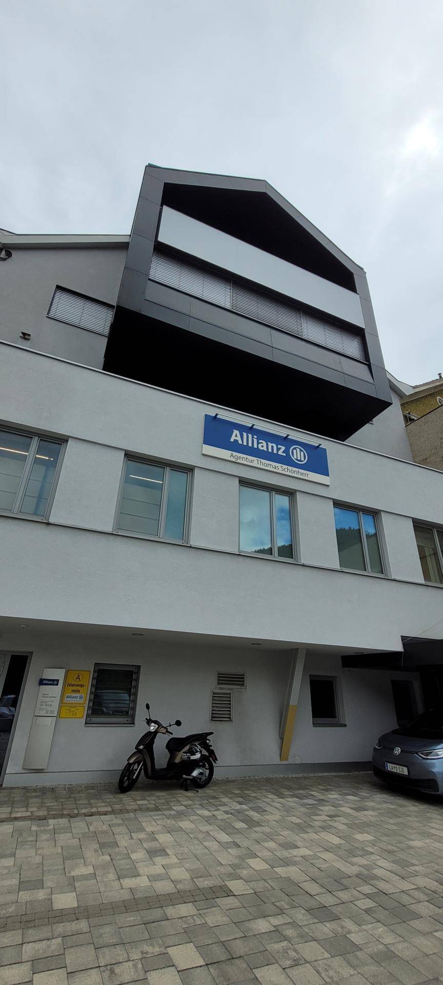 Moderne Gebäudefassade mit großen Fenstern, Allianz-Schild und gepflastertem Vorplatz.