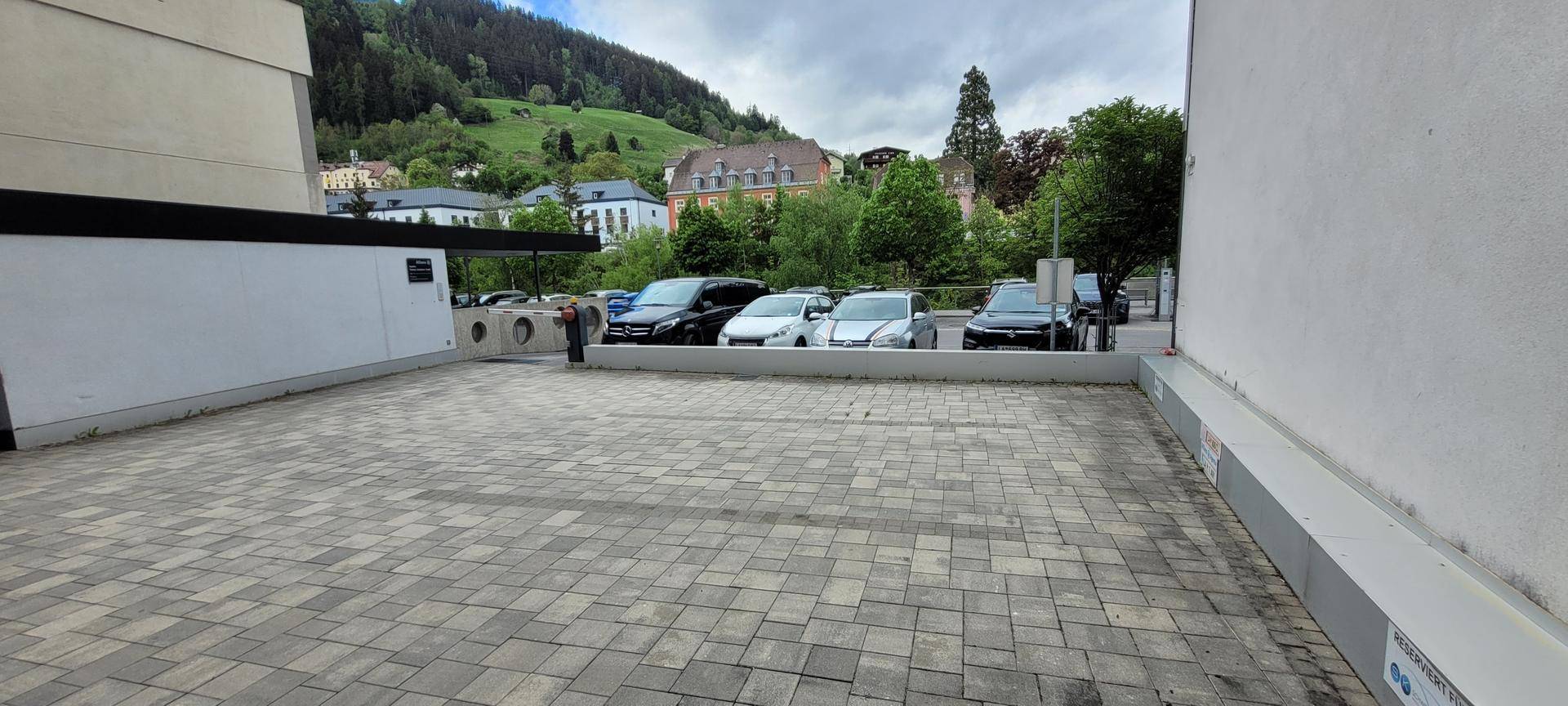 Großer gepflasterter Parkplatz mit Autos und Blick auf grüne Hügel im Hintergrund.