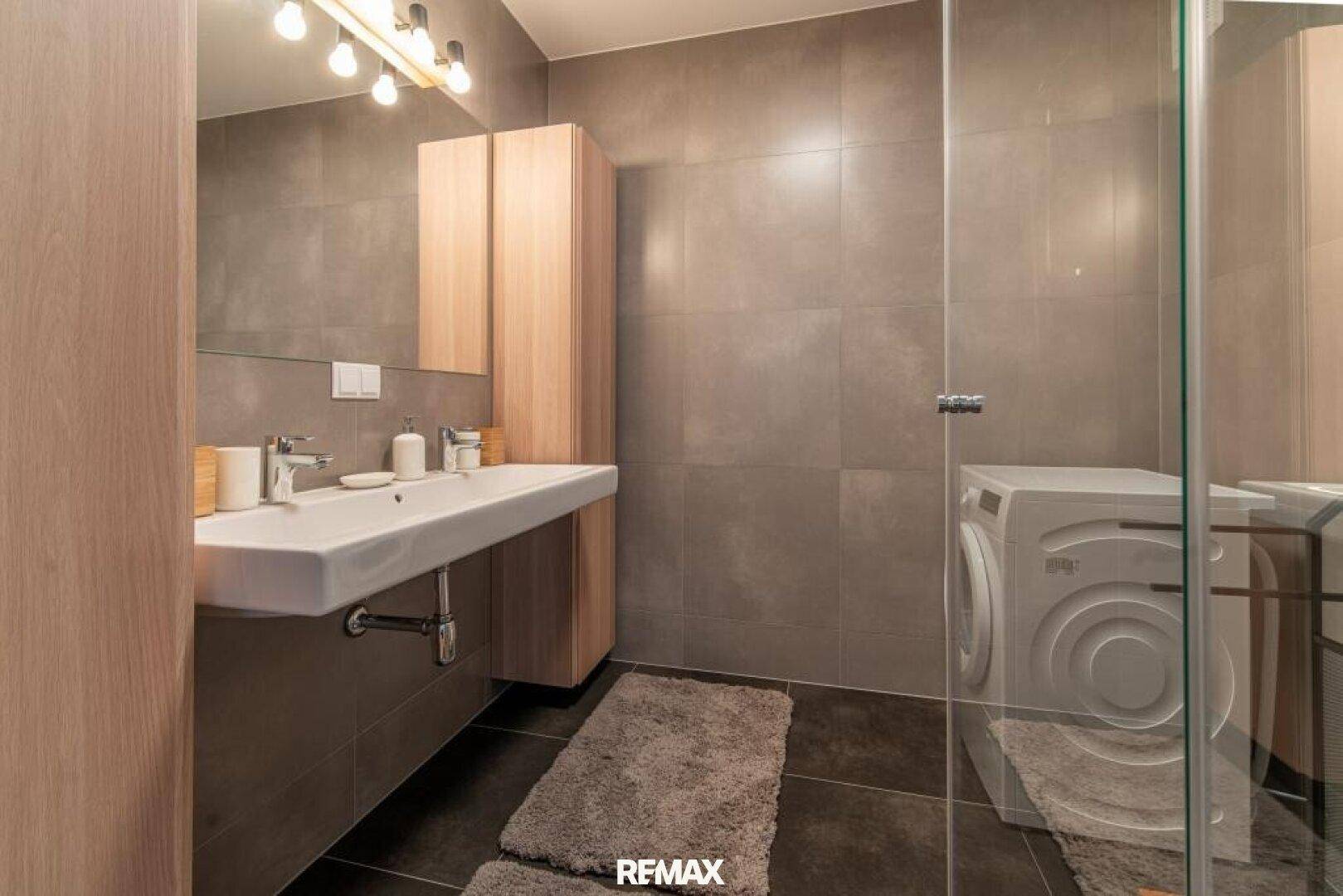 Modernes Badezimmer mit großformatigen grauen Fliesen, Waschbecken und Duschbereich.