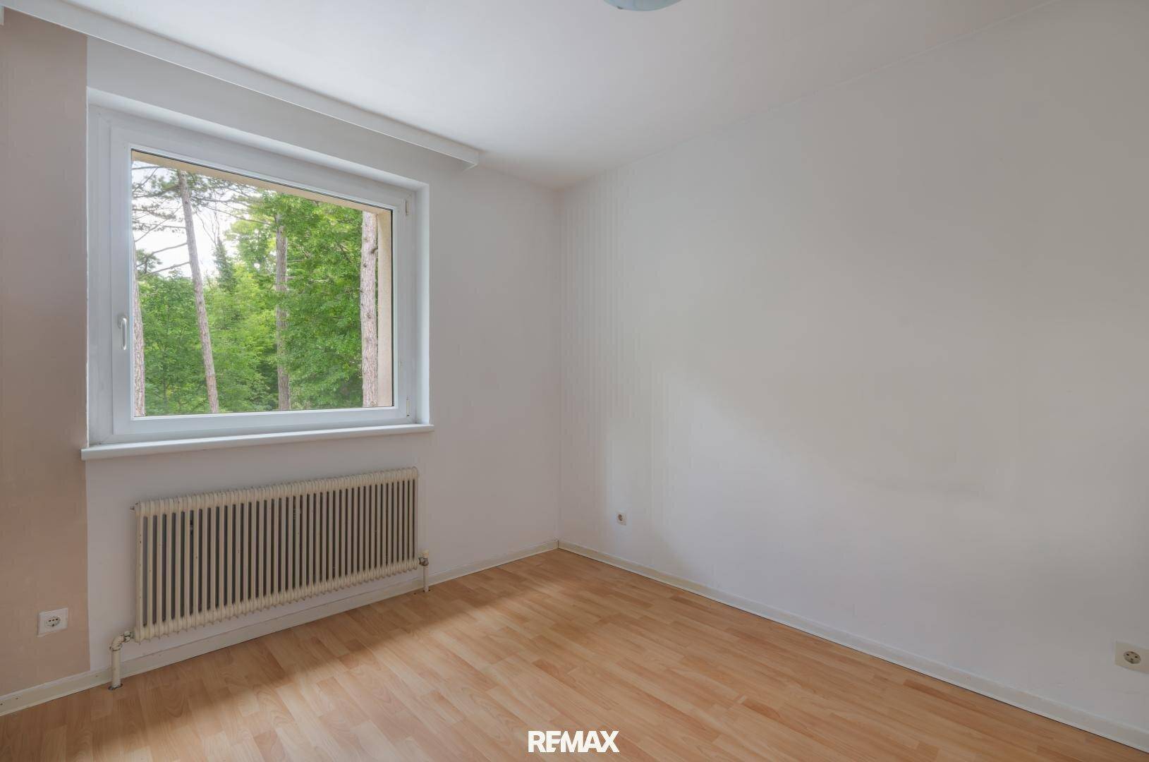 Leeres Schlafzimmer mit Parkettboden und großem Fenster mit Blick ins Grüne.