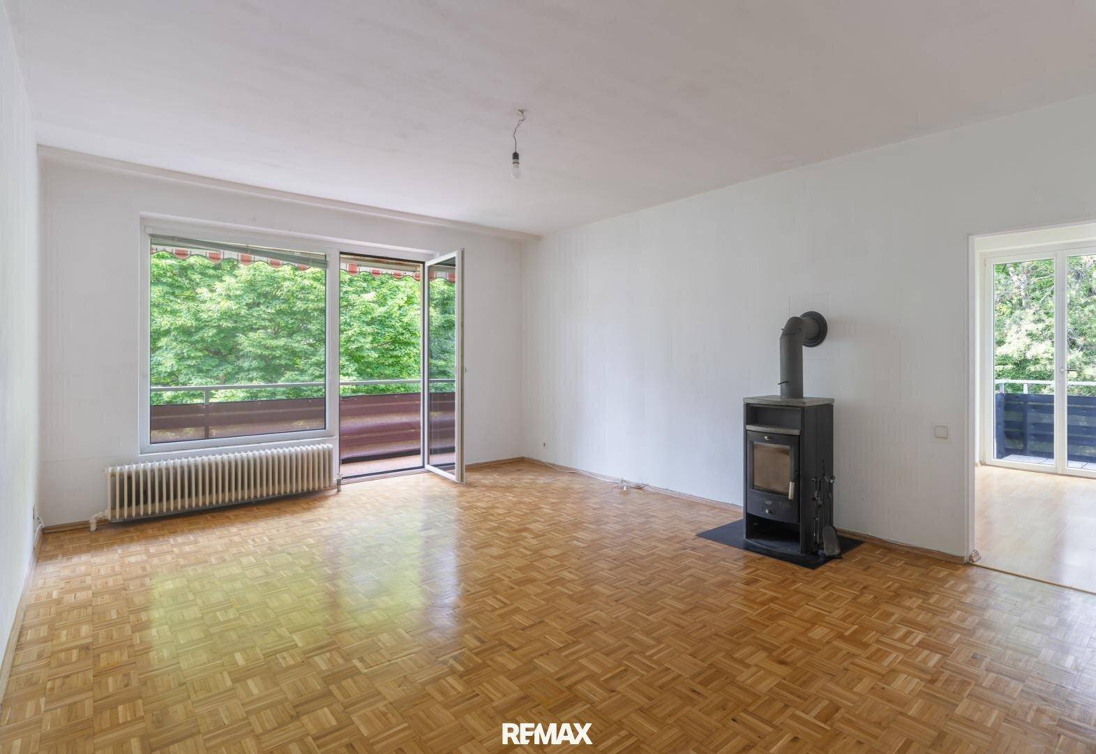Geräumiges, unmöbliertes Wohnzimmer mit Parkettboden, Kaminofen und Balkonzugang.