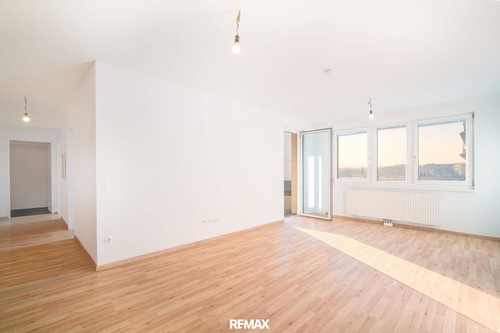 Leeres Wohnzimmer mit Holzboden, großen Fenstern und Zugang zum Balkon.
