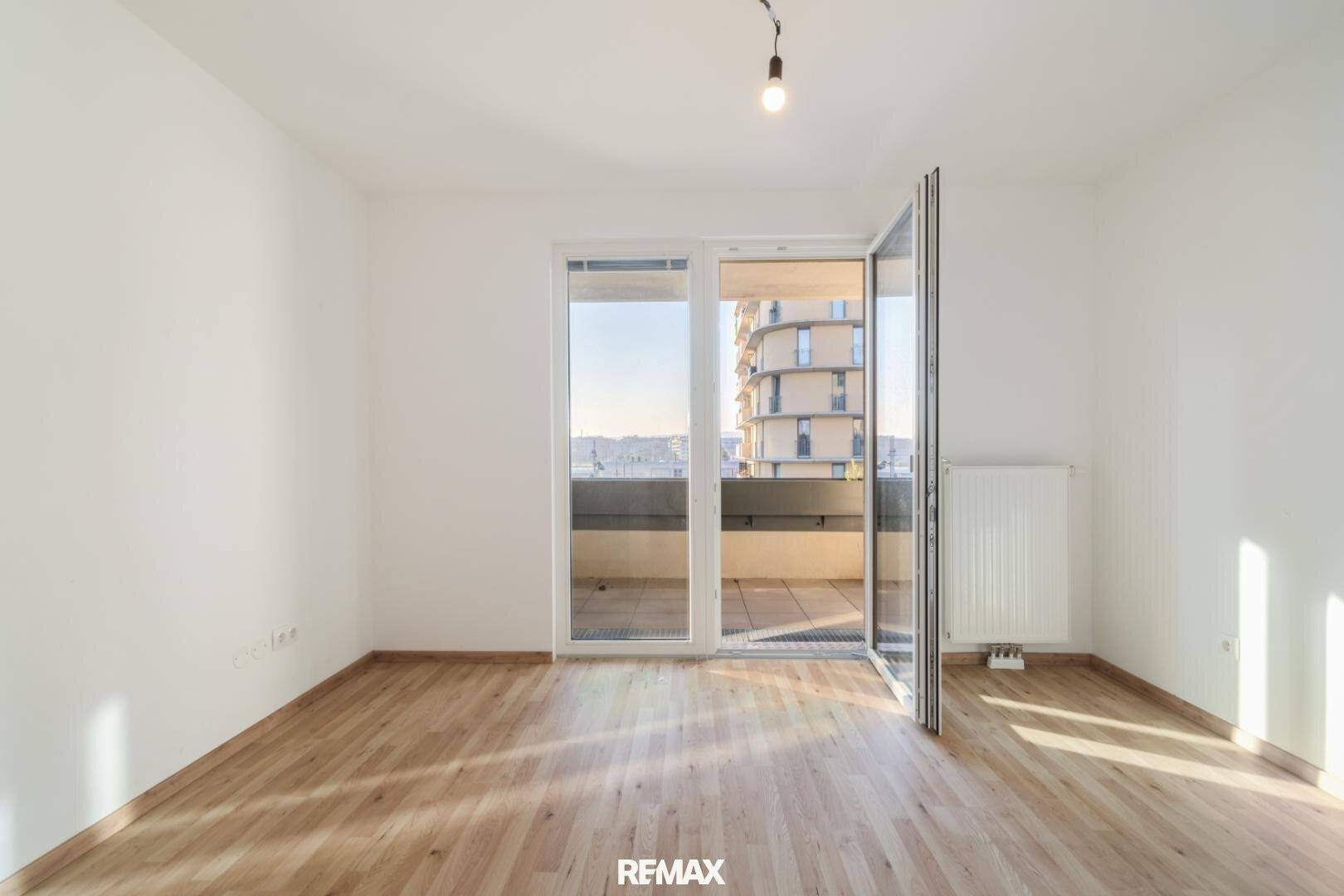 Leerer Raum mit Holzboden, großen Fenstern und Zugang zu einem Balkon.