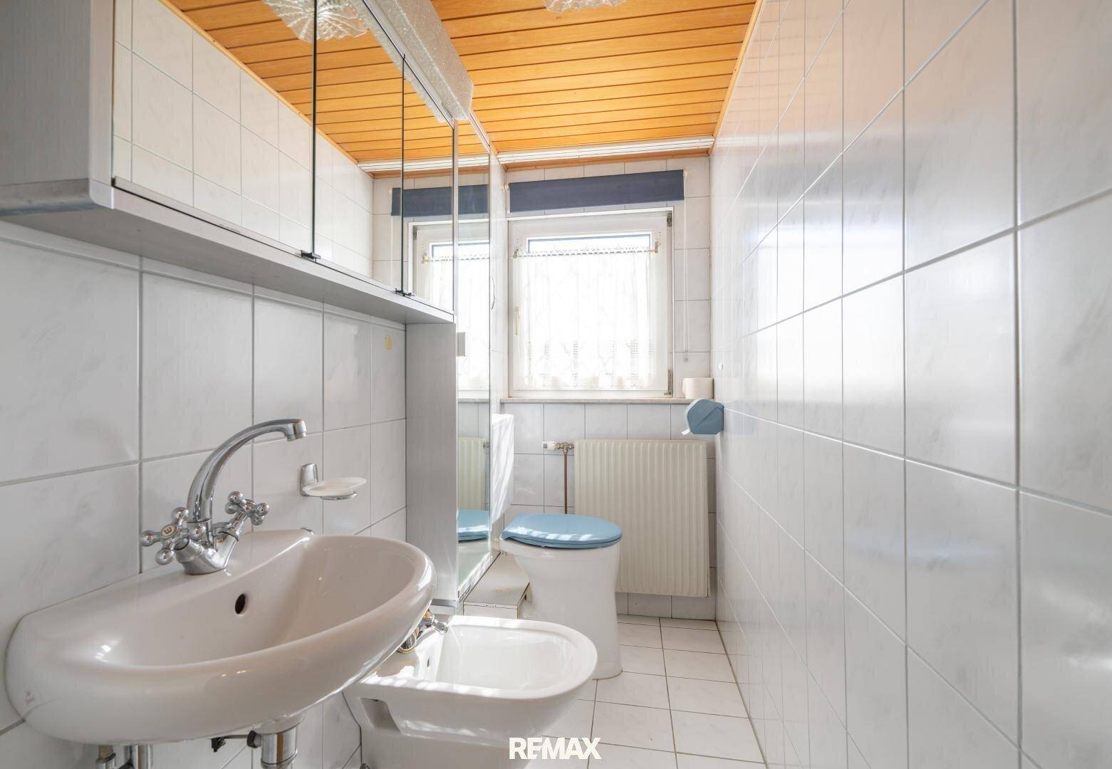 Kleines Badezimmer mit weißen Fliesen, Holzdecke, WC, Bidet und einem Fenster.