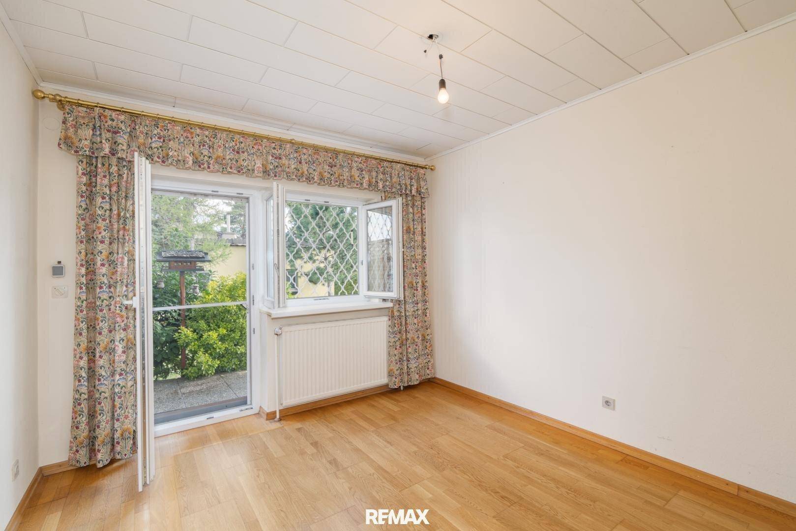 Helles Zimmer mit Holzfußboden und Zugang zu einem grünen Außenbereich, geschmückt mit Vorhängen.