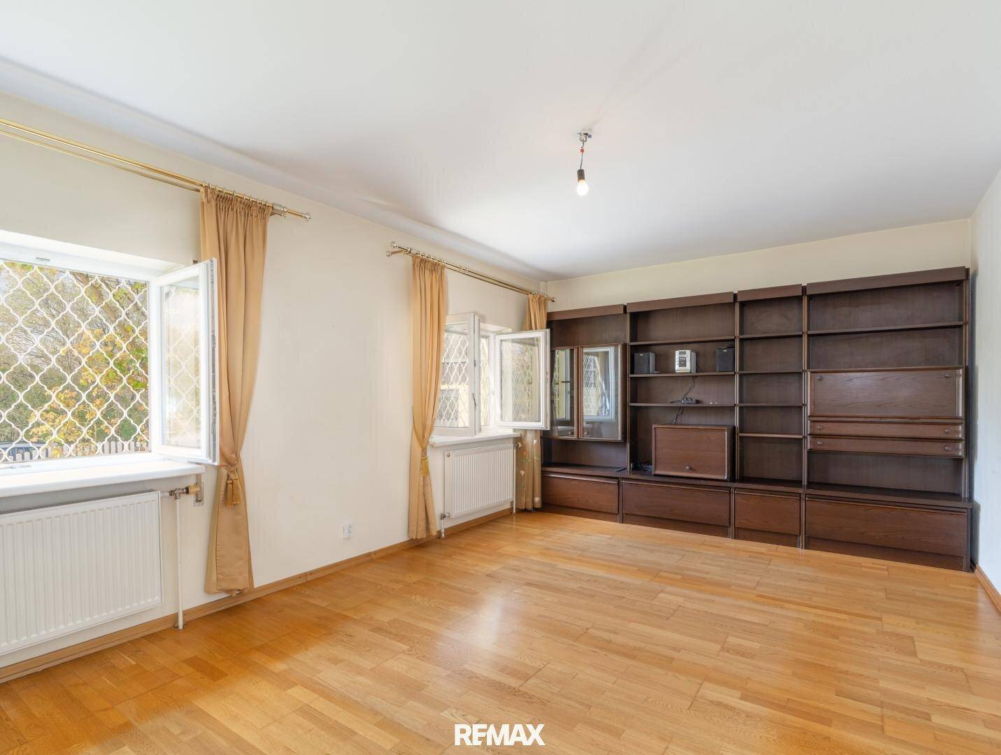 Helles Wohnzimmer mit Holzfußboden, zwei Fenstern und einer großen, dunklen Holzregalwand.