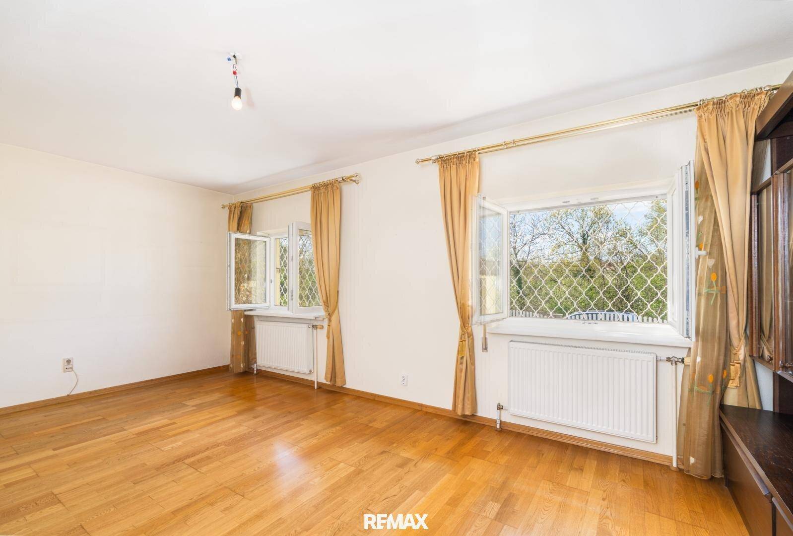 Geräumiges Wohnzimmer mit hellem Holzfußboden und zwei großen Fenstern, die viel Licht hereinlassen.
