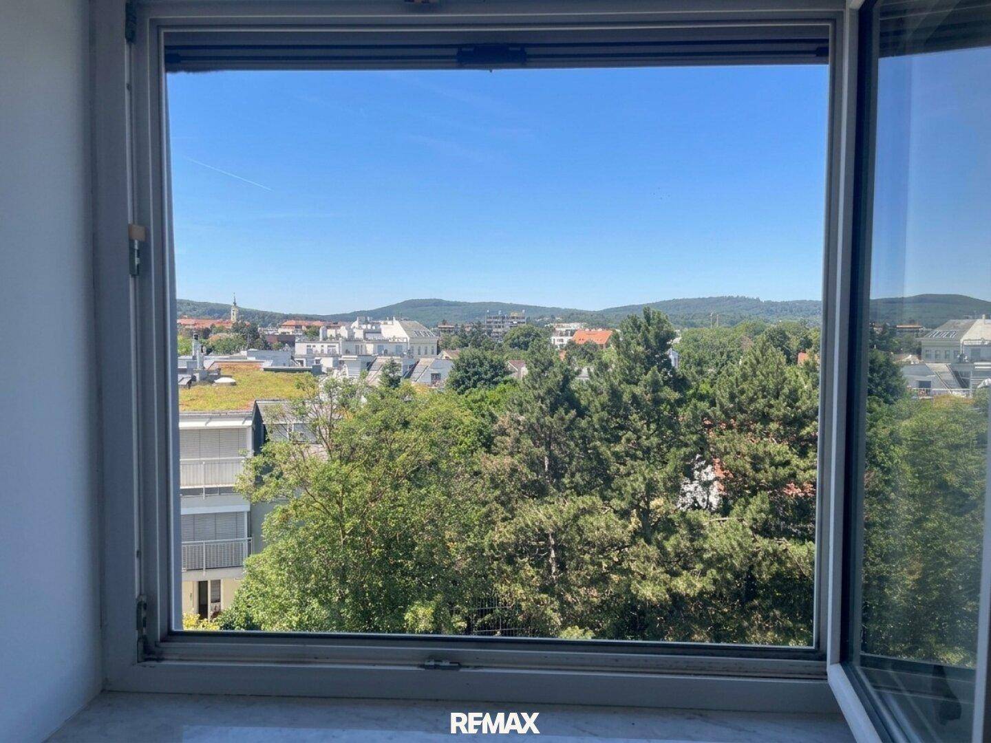 Heller Ausblick aus einem Fenster auf grüne Bäume, Gebäude und eine hügelige Landschaft.