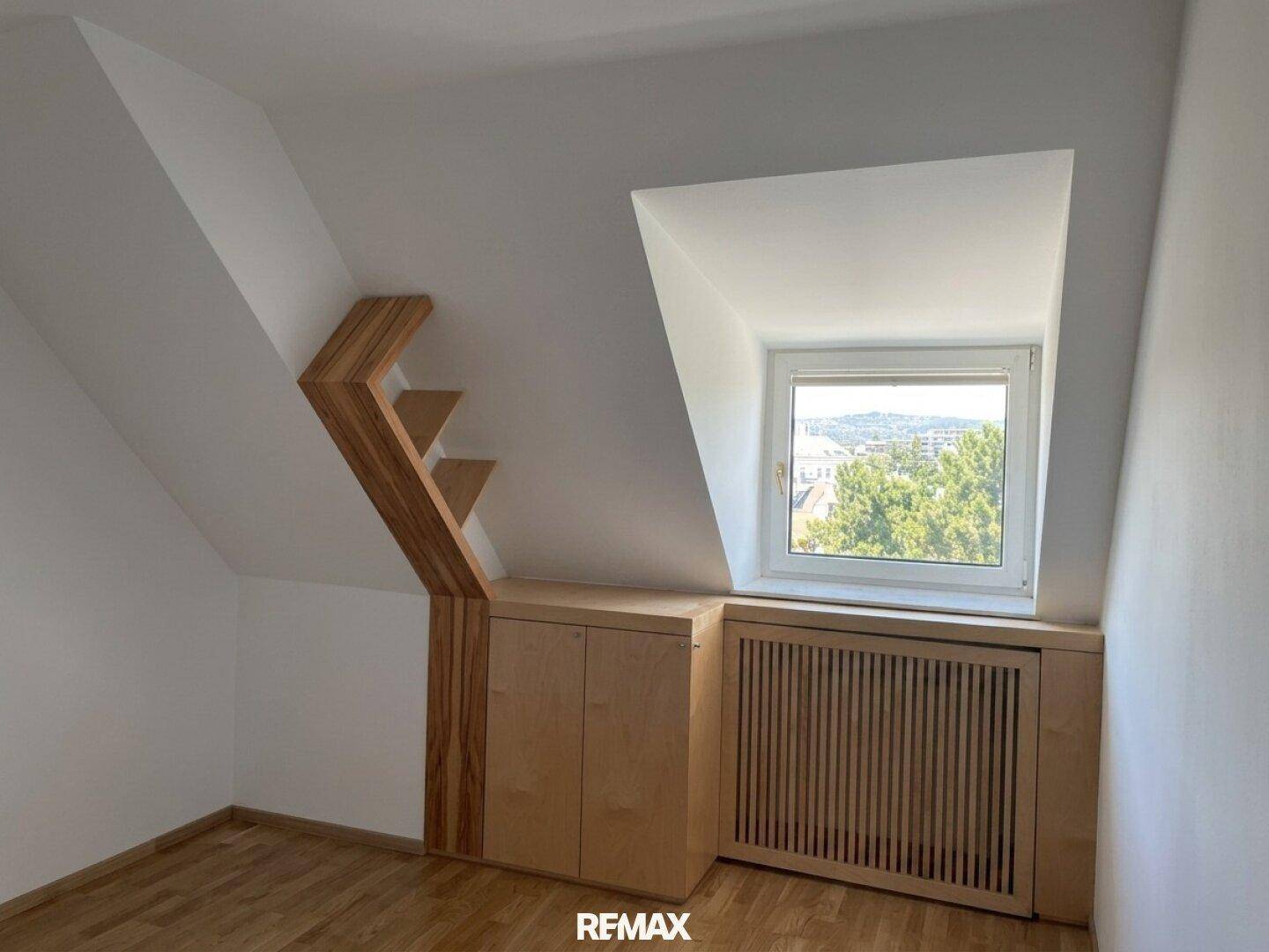 Dachzimmer mit schrägen Wänden, Holzeinbauten und einem Fenster mit Ausblick.