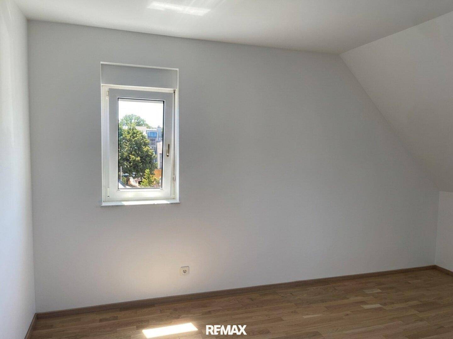 Helles Schlafzimmer mit Holzboden, weiß gestrichenen Wänden und Fenster, unmöbliert.