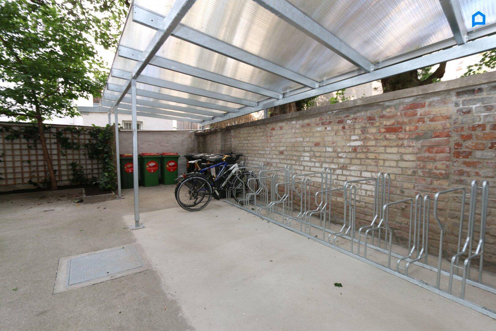Überdachter Fahrradabstellplatz mit Fahrradständern und Mülltonnen im Außenbereich des Gebäudes.