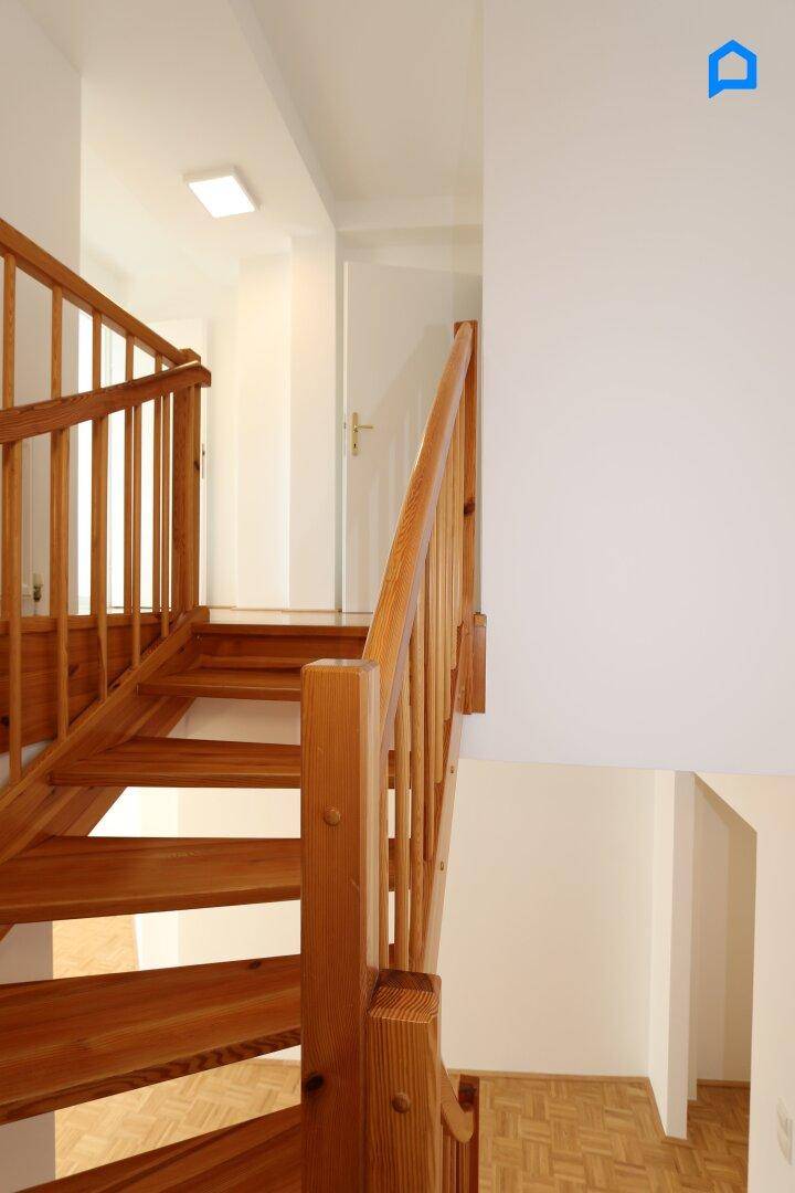 Holztreppe führt zu einem hellen Flur mit weiß gestrichenen Wänden und Deckenleuchte.
