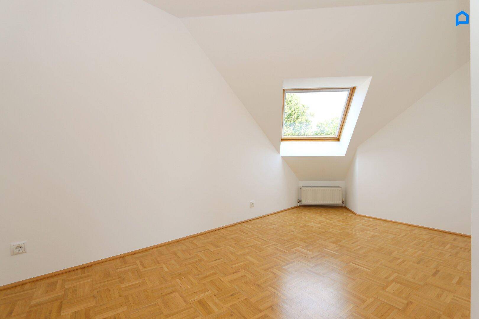 Helles Zimmer im Dachgeschoss mit Parkettboden und einem Dachfenster, das viel Tageslicht hereinlässt.