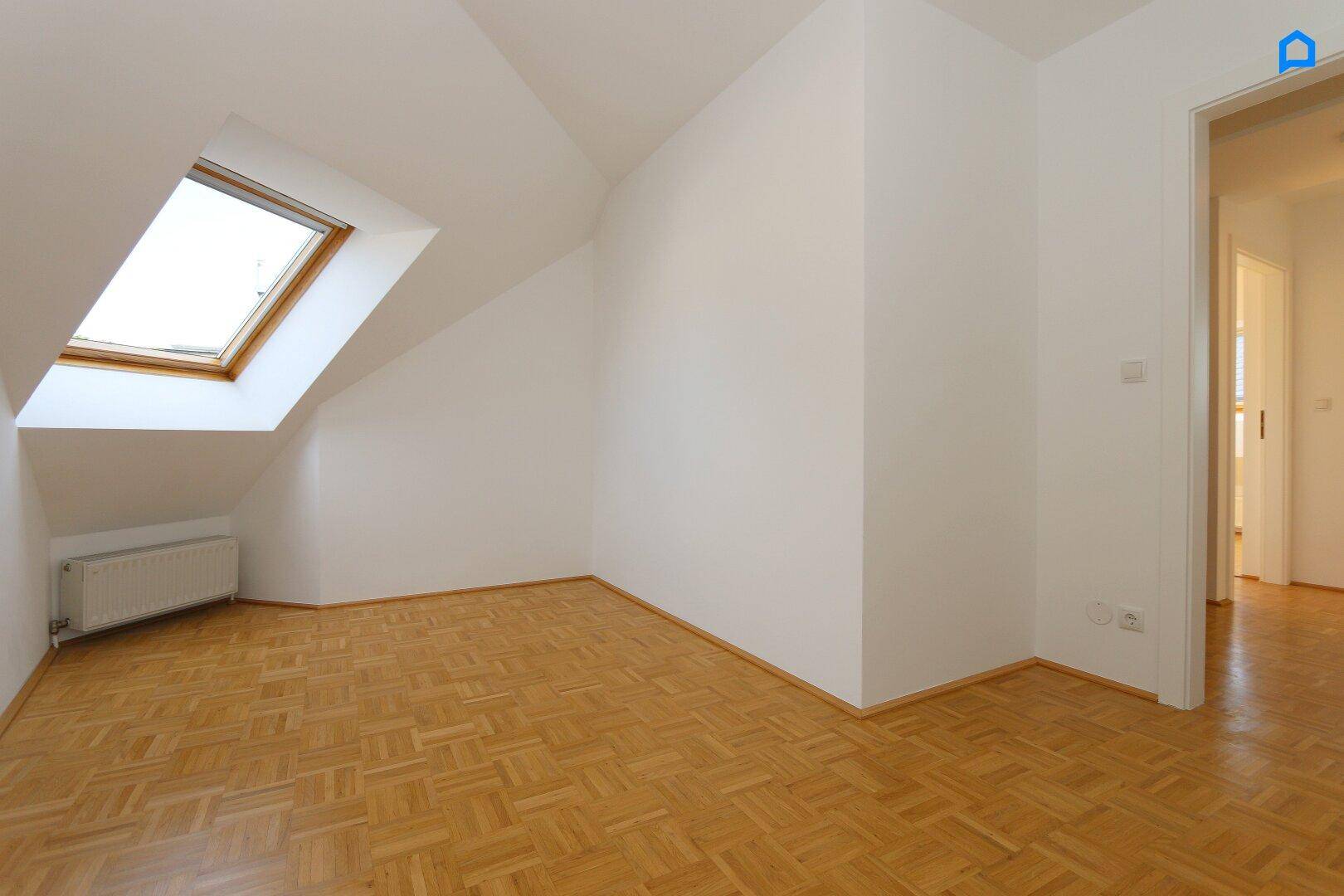 Helles Zimmer im Dachgeschoss mit Parkettboden und einem Dachfenster, das viel Tageslicht hereinlässt.
