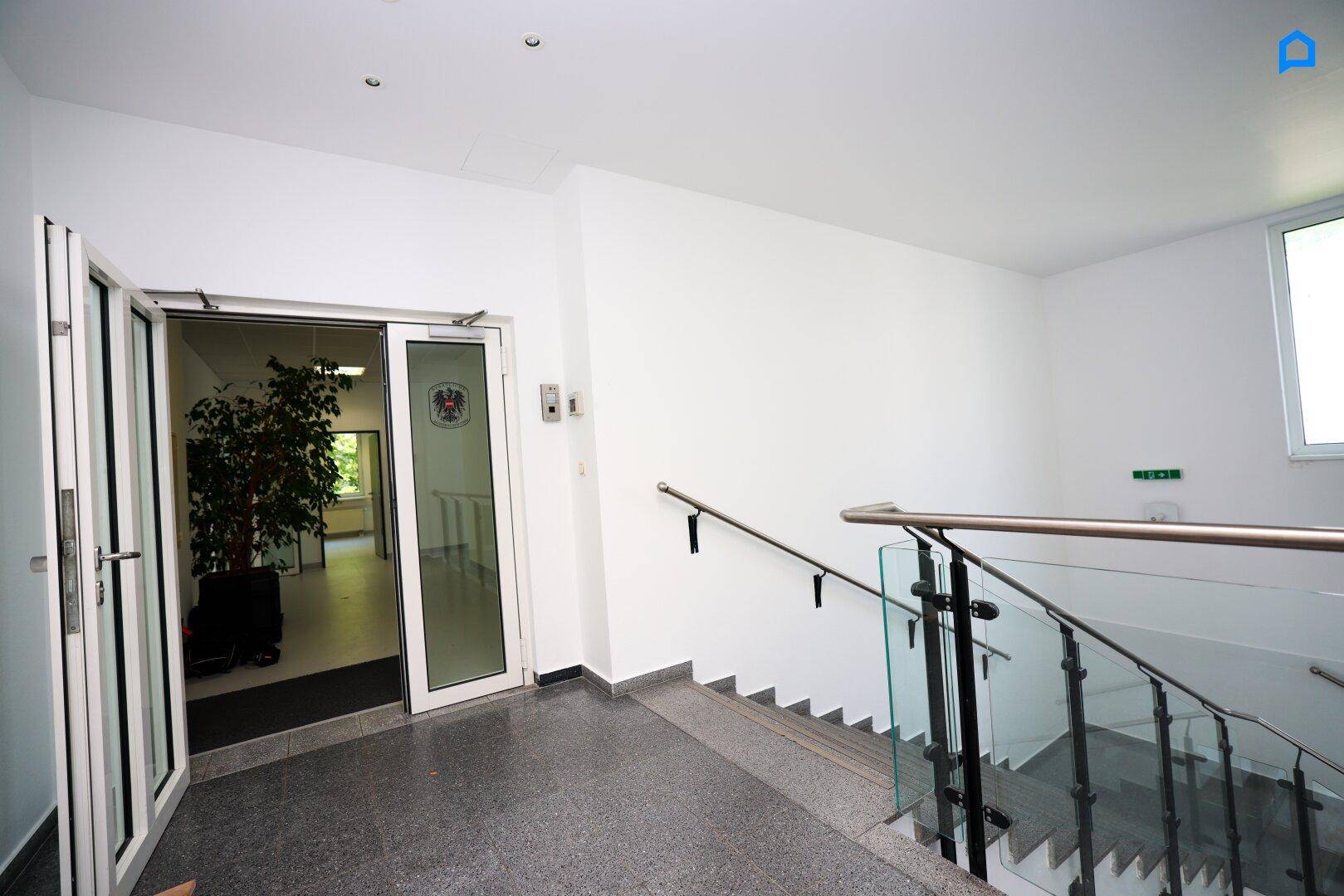Eingangsbereich mit Treppe, Glasgeländer und doppelten Glastüren zum Gebäude.