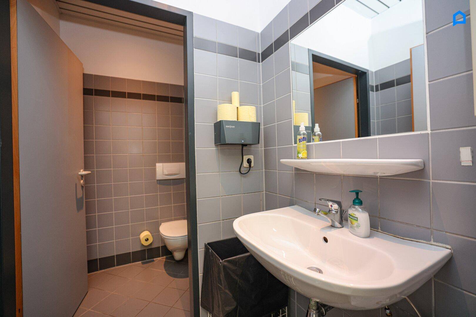 Badezimmer mit grauen Fliesen, Waschbecken, Spiegel und Toilette.