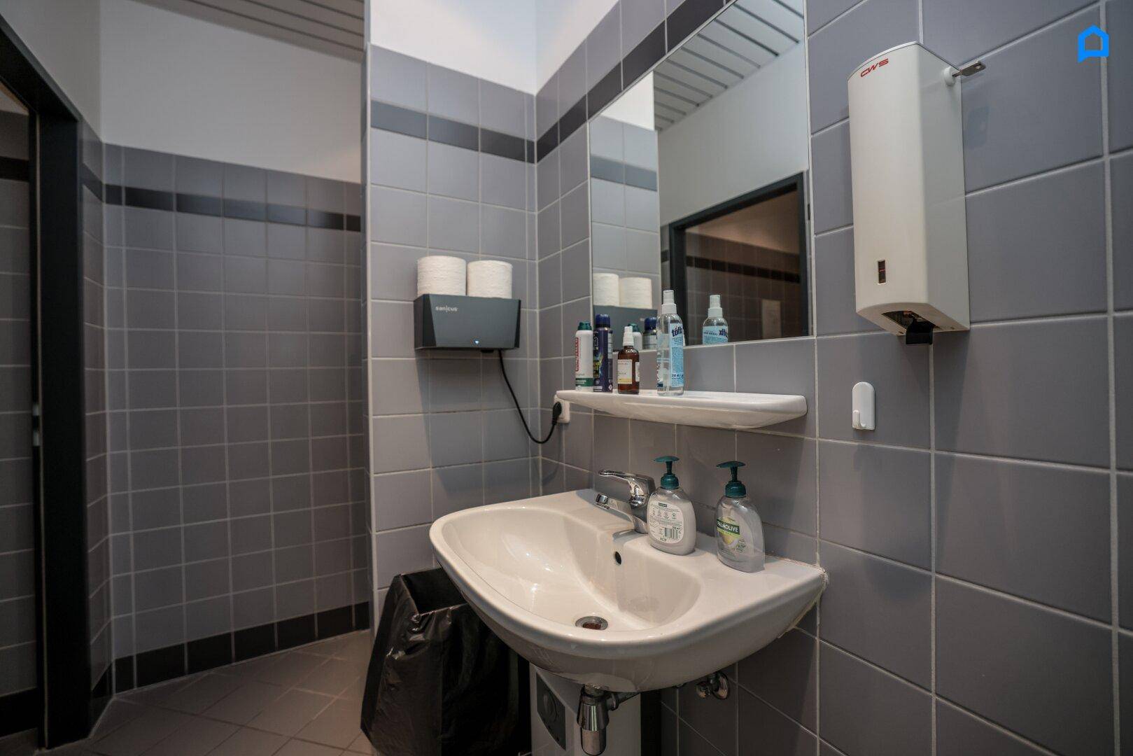 Badezimmer mit grauen Fliesen an Wänden und Boden, Waschbecken und Spiegel.