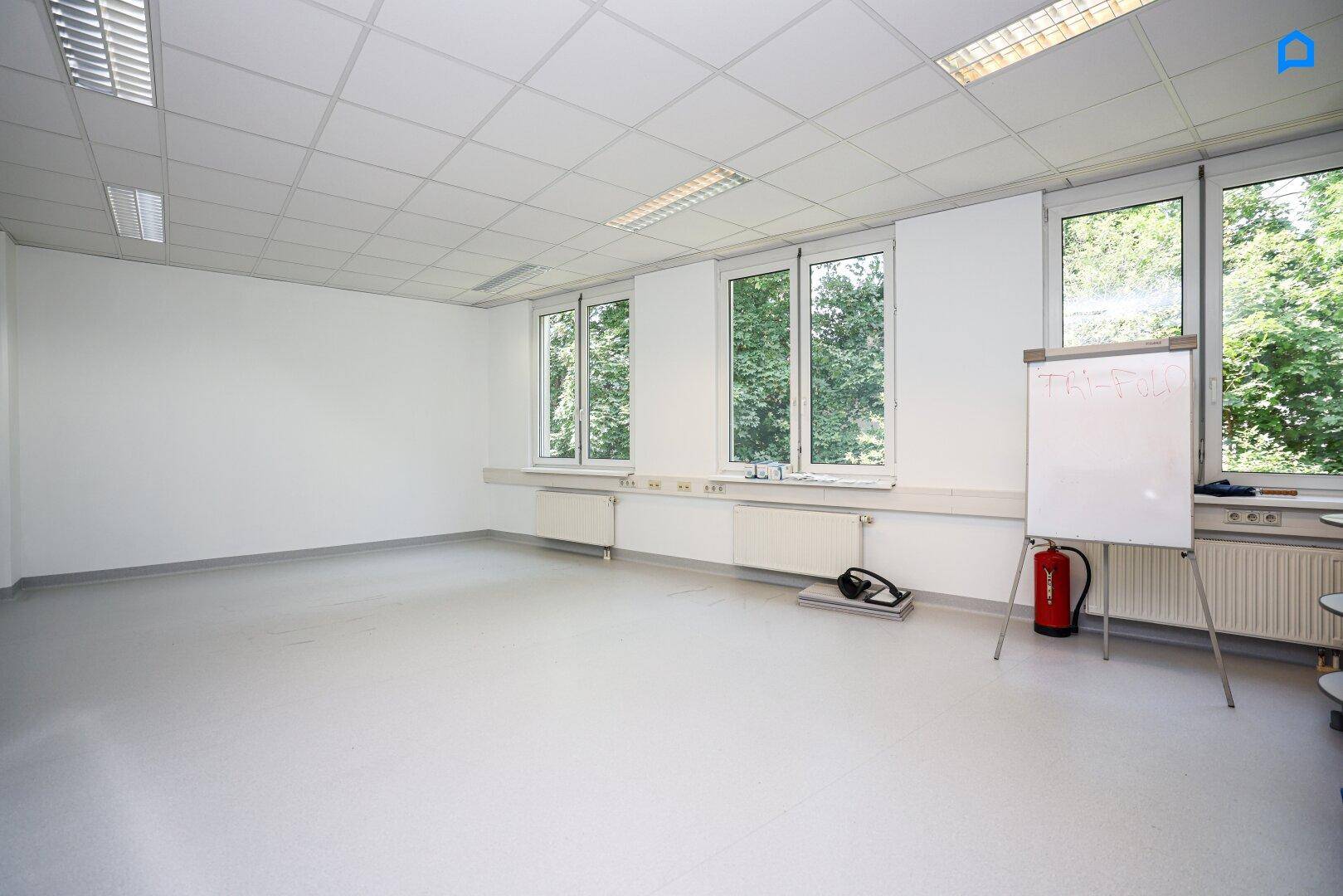 Heller, leerer Raum mit großen Fenstern, Whiteboard und Feuerlöscher.