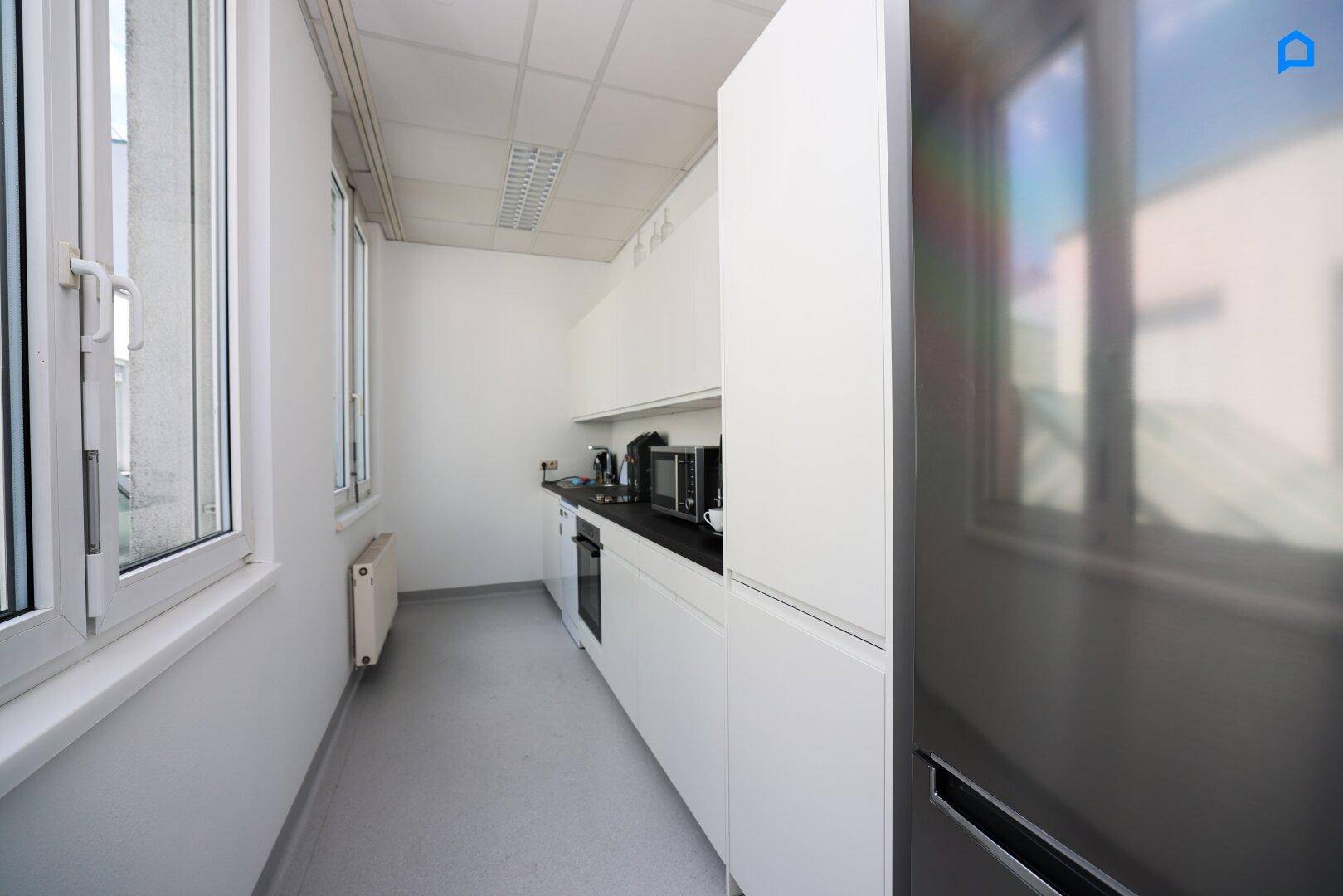 Schmale, moderne Teeküche mit weißen Schränken, dunkler Arbeitsplatte und großem Kühlschrank.