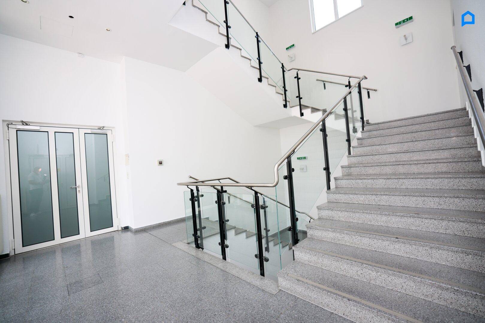 Modernes Treppenhaus mit Glasgeländer, Edelstahlhandläufen und grauem Fliesenboden.