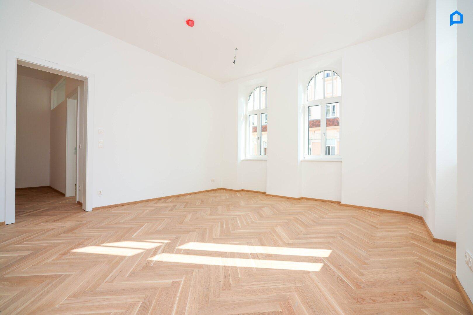 Helles Zimmer mit Fischgrätparkett und sonnigen Lichtstreifen durch die Rundbogenfenster.