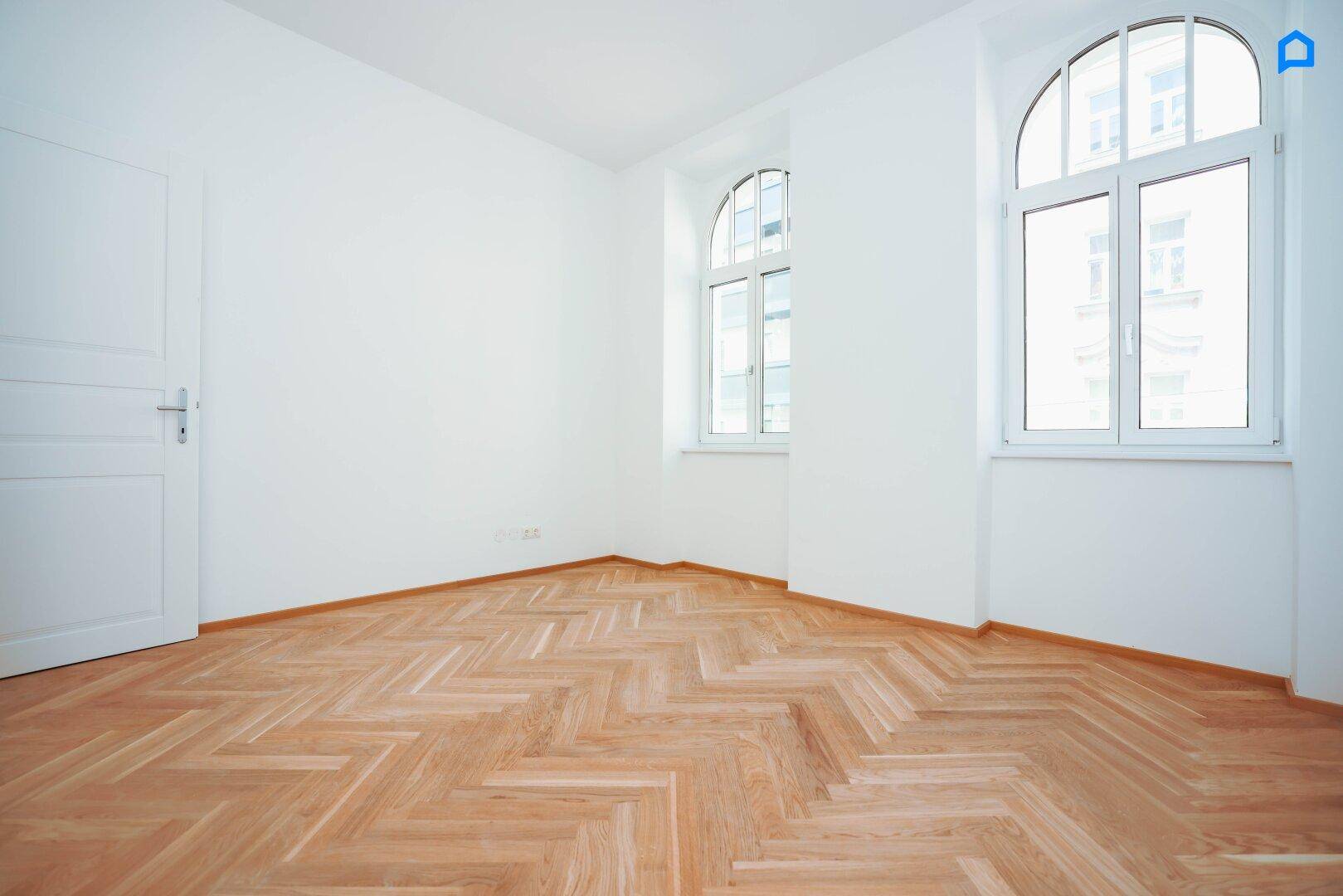 Lichtdurchflutetes Zimmer mit Fischgrätparkett und hohen Rundbogenfenstern, sehr hell.