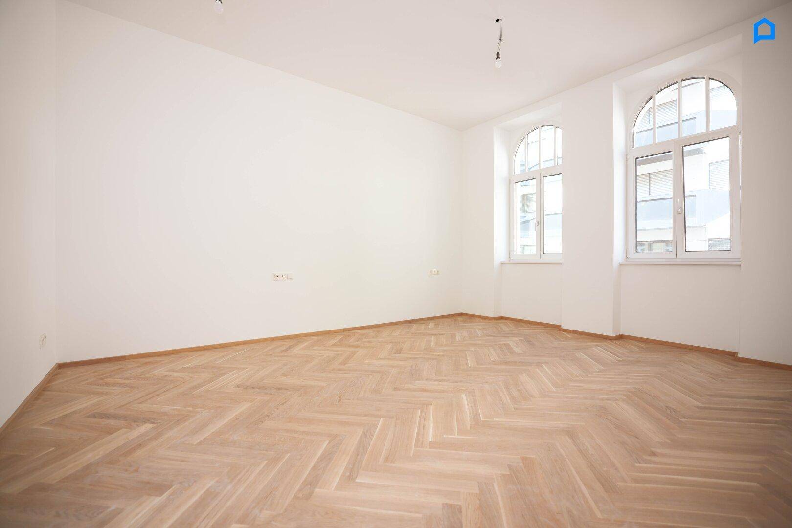 Geräumiges Zimmer mit Fischgrätparkett und zwei großen Rundbogenfenstern, lichtdurchflutet.
