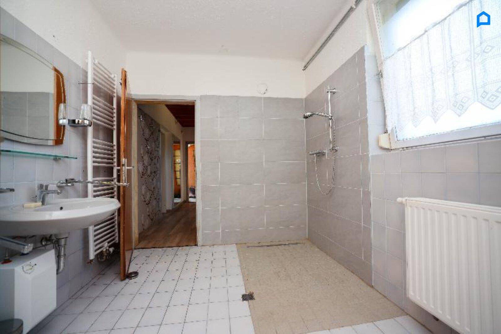 Badezimmer mit Dusche, Waschbecken, Heizkörper und Fenster, hell gefliest.