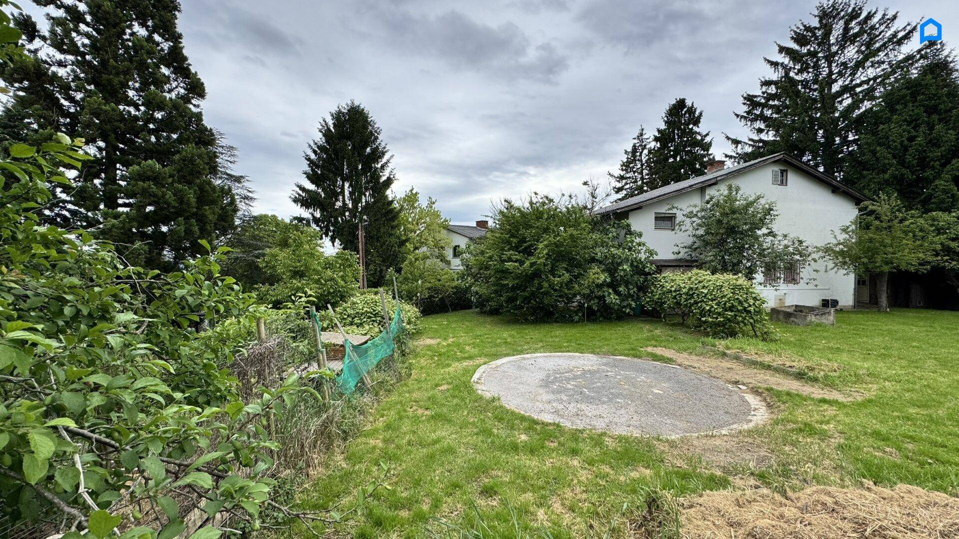 Gartenbereich mit Rasen, Bäumen und einer runden gepflasterten Fläche neben dem Haus.