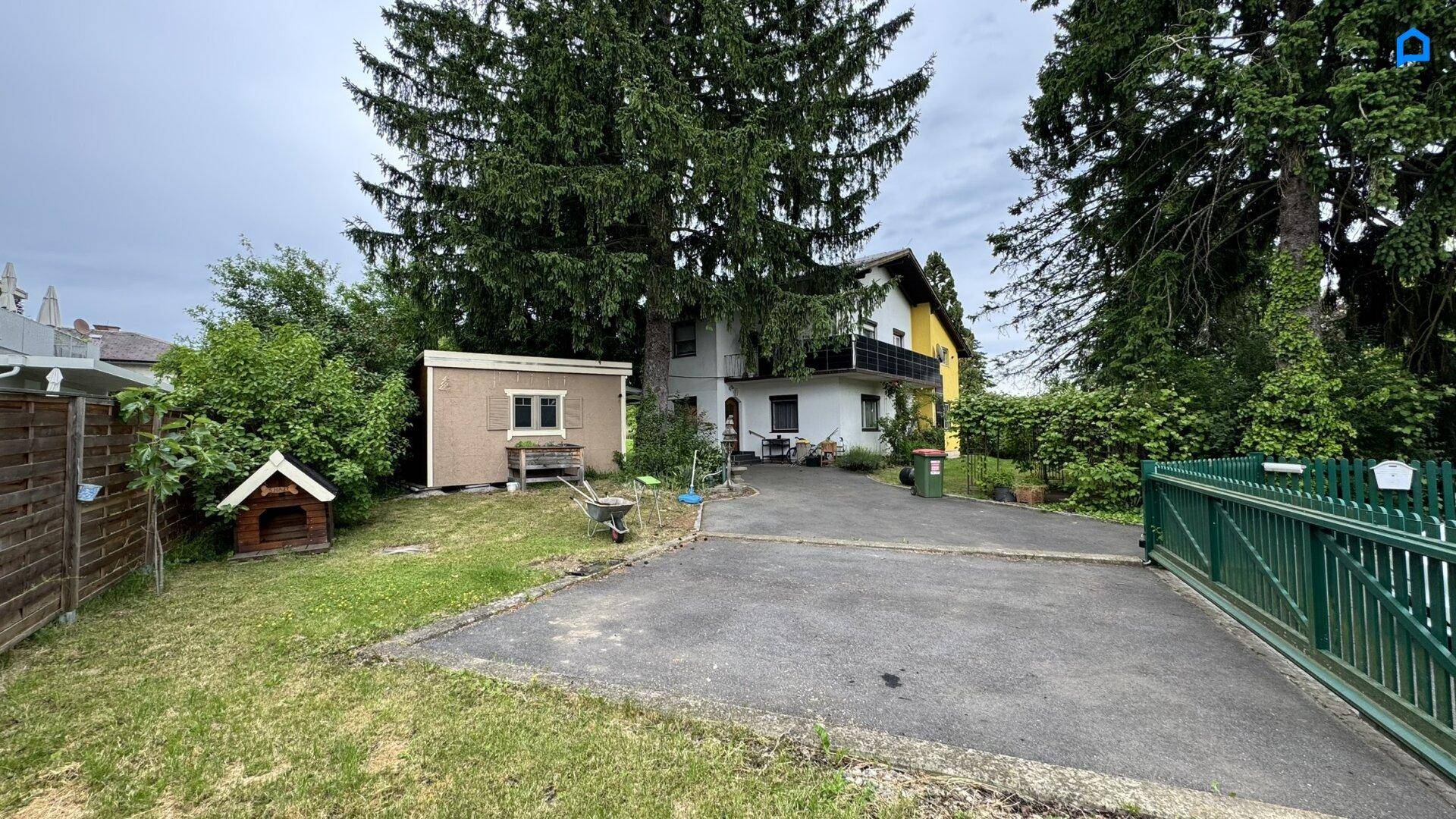 Vorderansicht des Hauses mit Zufahrt, Gartenhaus und umgebenden Bäumen.