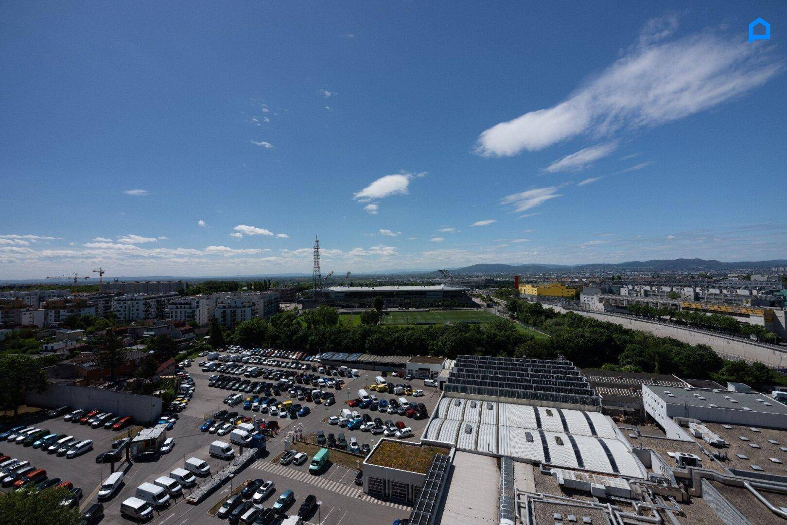 Weitläufiger Blick über die Stadt mit Parkplätzen und einem Stadion im Hintergrund.