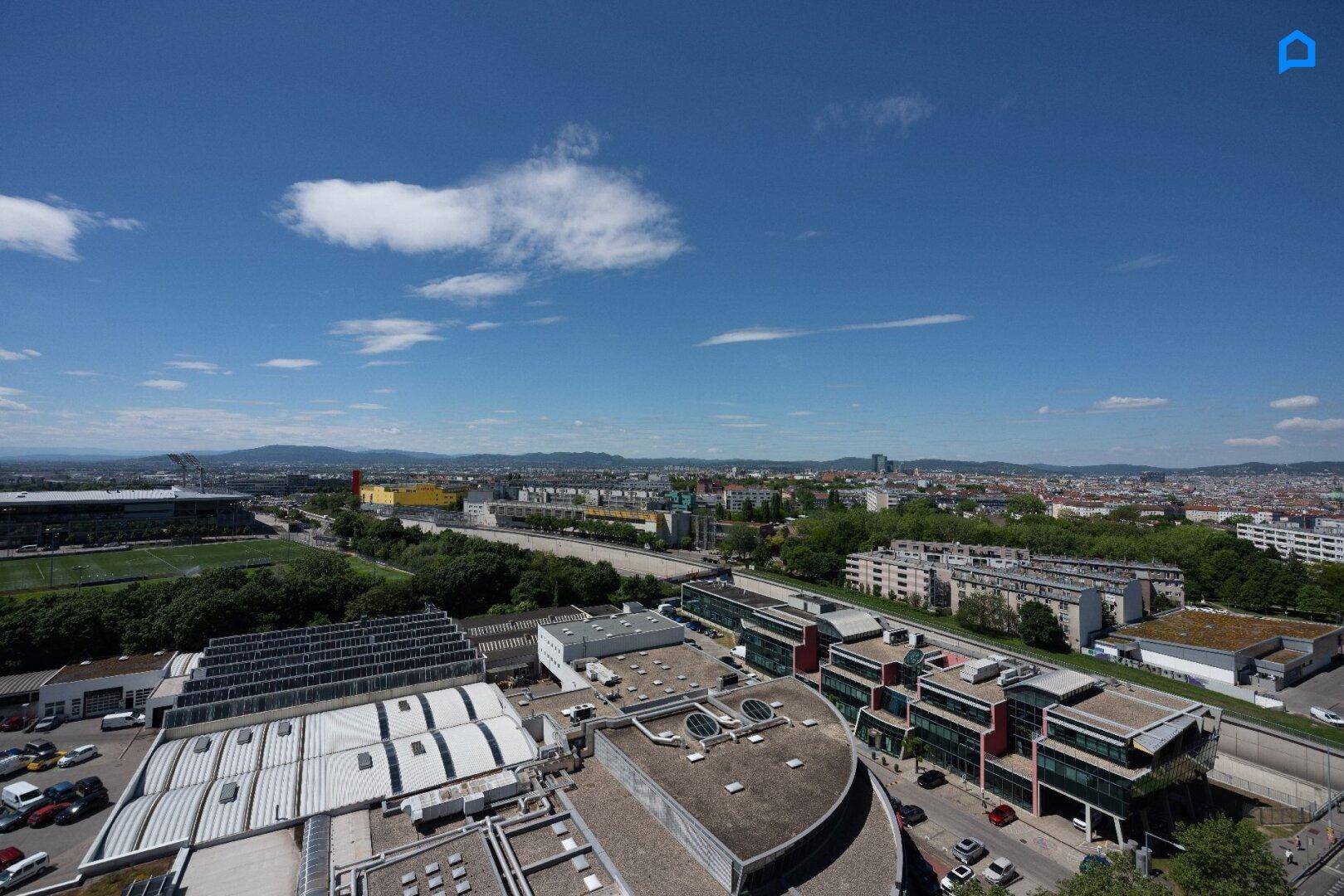 Panoramablick über die Stadtlandschaft mit Gebäuden, Straßen und grünen Hügeln am Horizont.