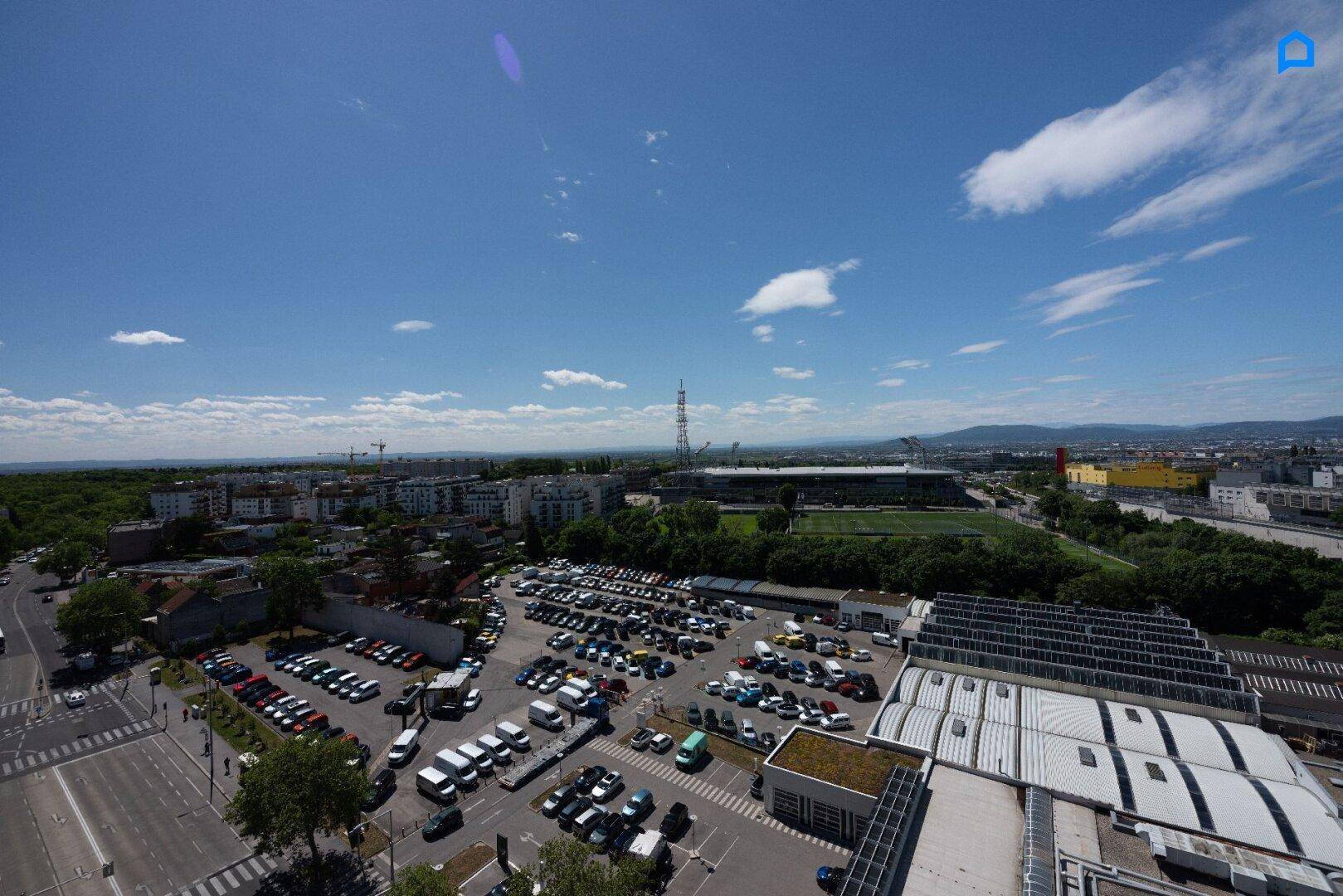 Weitläufiger Blick über die Stadt mit Parkplätzen und einem Stadion im Hintergrund unter blauem Himmel.