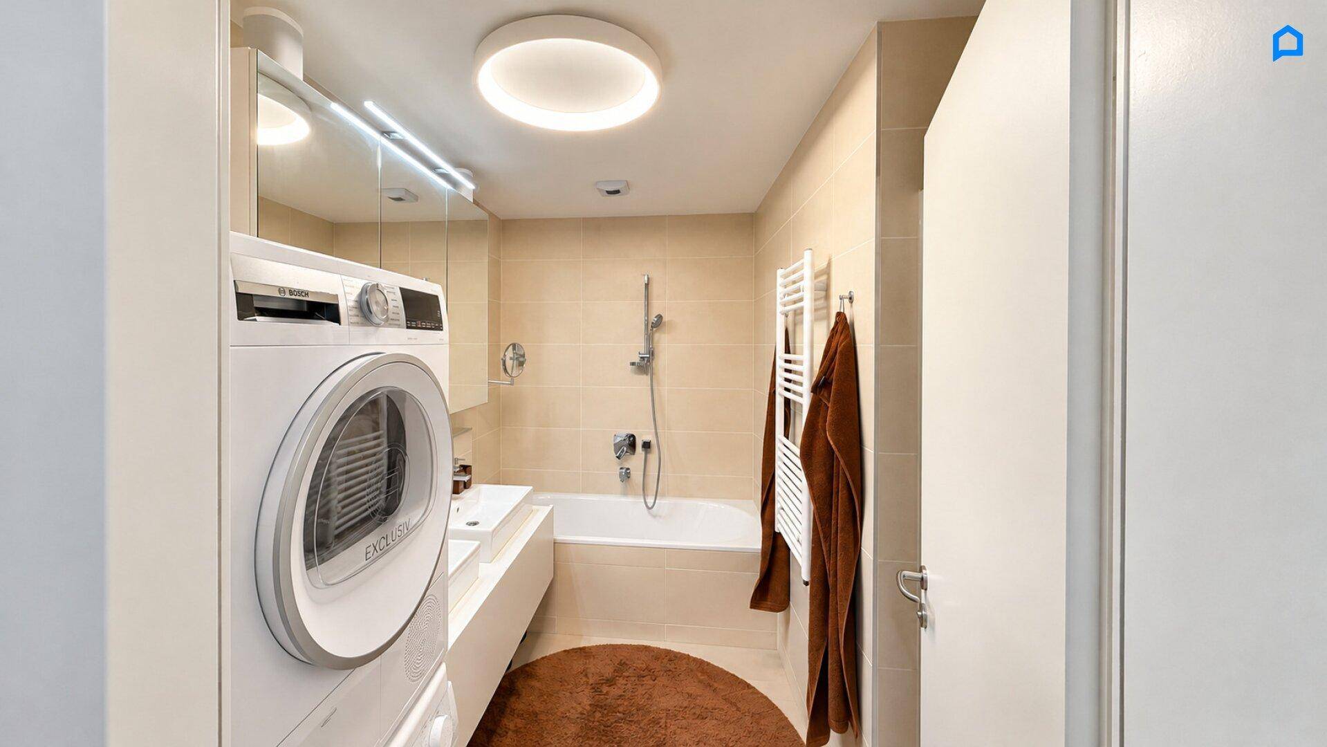 Modernes Badezimmer mit Badewanne, Waschmaschine und Trockner sowie beigefarbenen Fliesen.