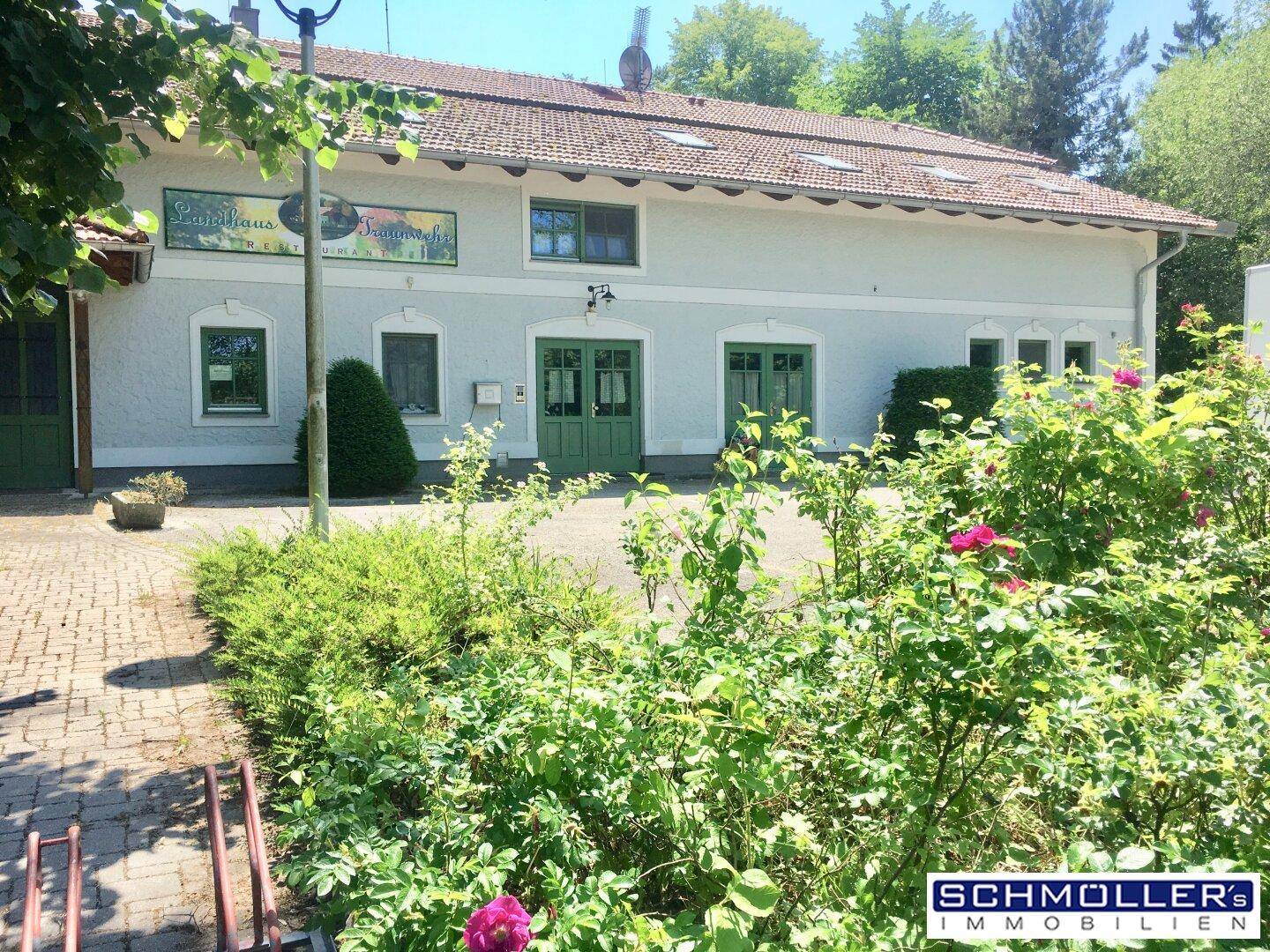Ansicht des Landhauses von vorne mit gepflastertem Bereich und grünen Büschen an einem sonnigen Tag.