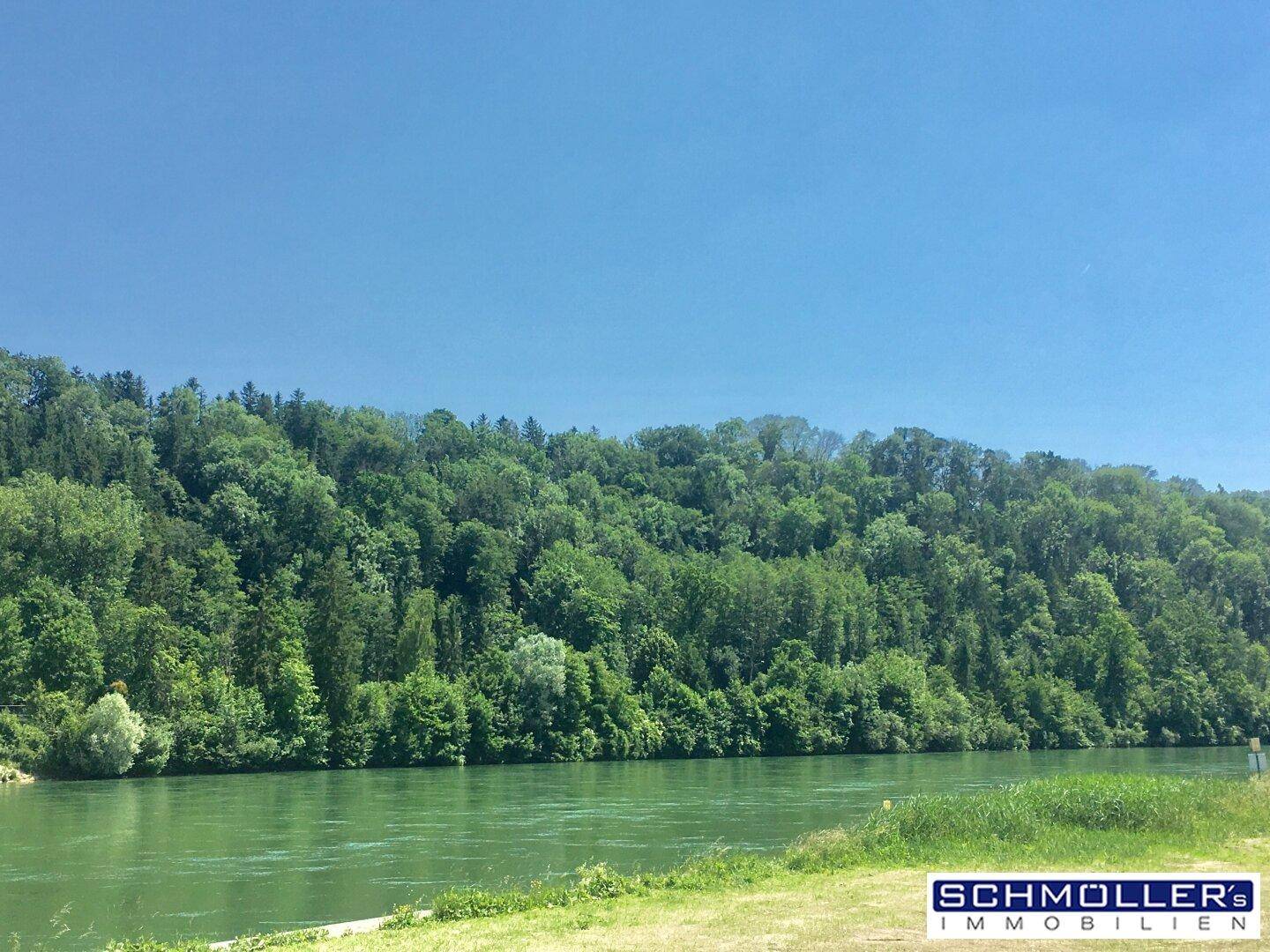 Breiter Fluss mit grünen Ufern und bewaldeten Hügeln unter strahlend blauem Himmel.